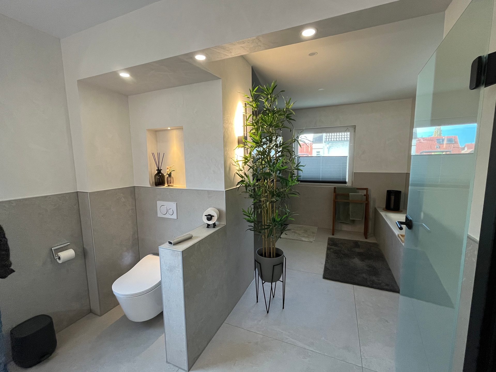 A contemporary bathroom with a toilet, a large potted plant, and a bathtub in neutral grey and white tones.
