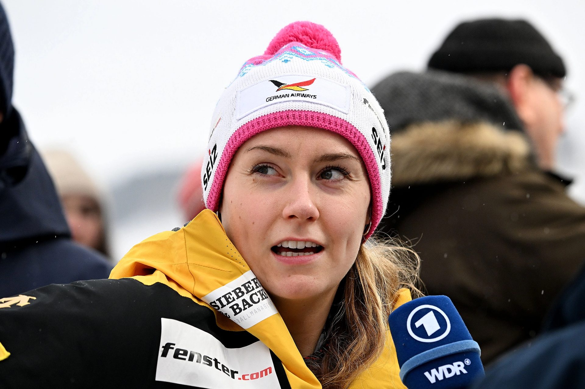 Woman in a white and pink "German Airways" hat talking, with a WDR microphone nearby.