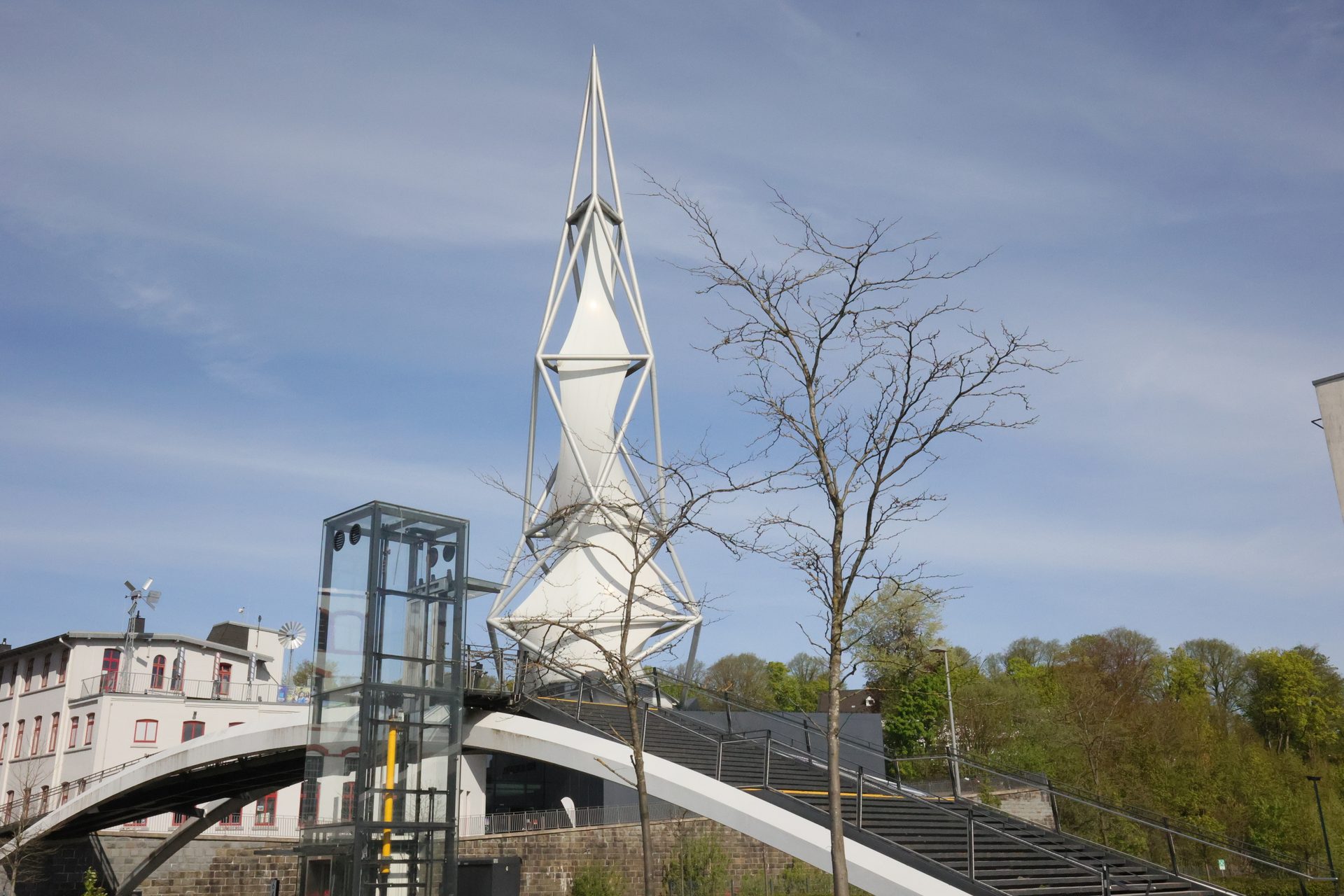 A modern white spire, glass elevator, and pedestrian bridge by buildings and trees under blue sky.