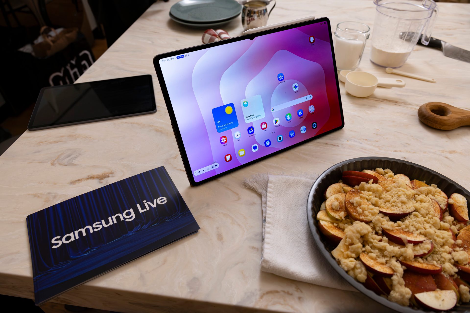 Samsung tablet displaying home screen, apple crumble, and "Samsung Live" booklet on a kitchen counter.