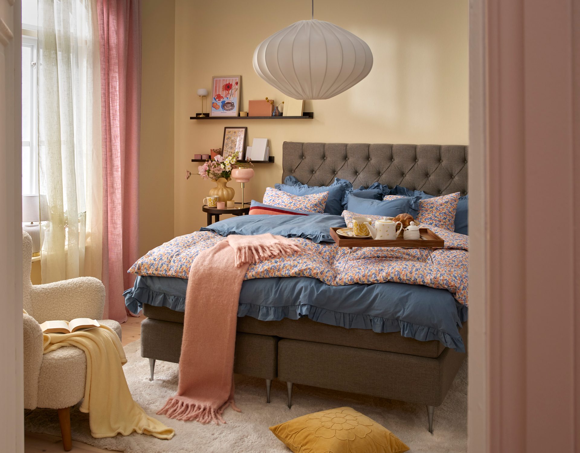 A cozy bedroom with a bed featuring blue and floral bedding, a breakfast tray, pink throw, and an armchair by sheer curtains.