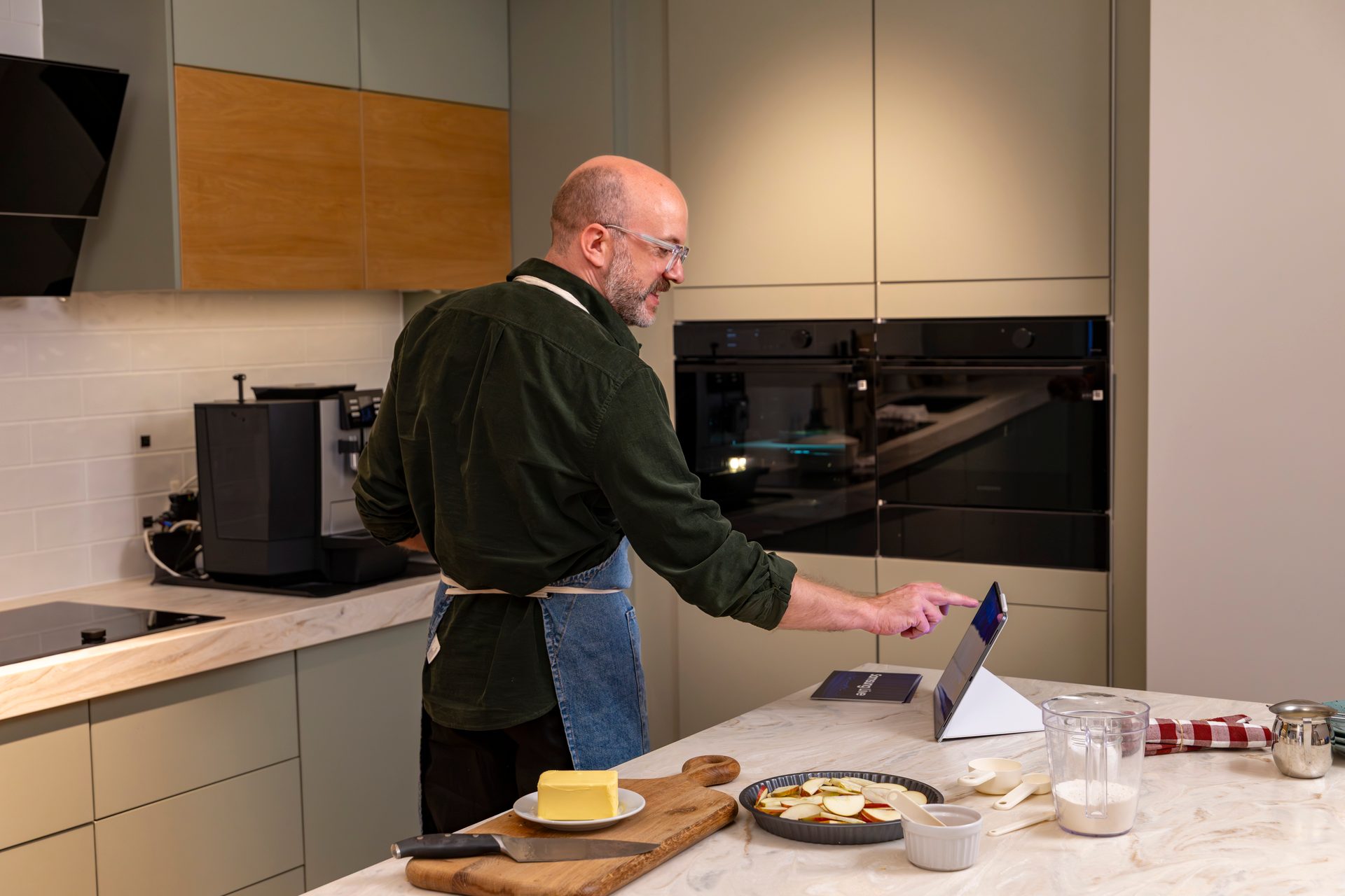 A man in a modern kitchen uses a tablet for a recipe, with ingredients like butter and apples laid out.