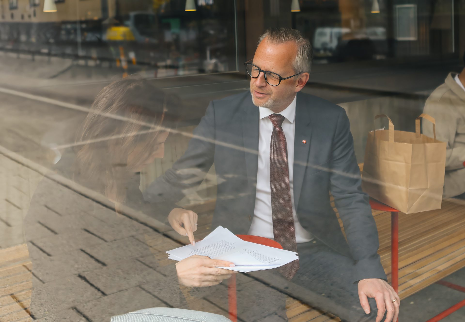 Two people, a man in a suit and a woman, discuss documents, viewed through a reflective window.