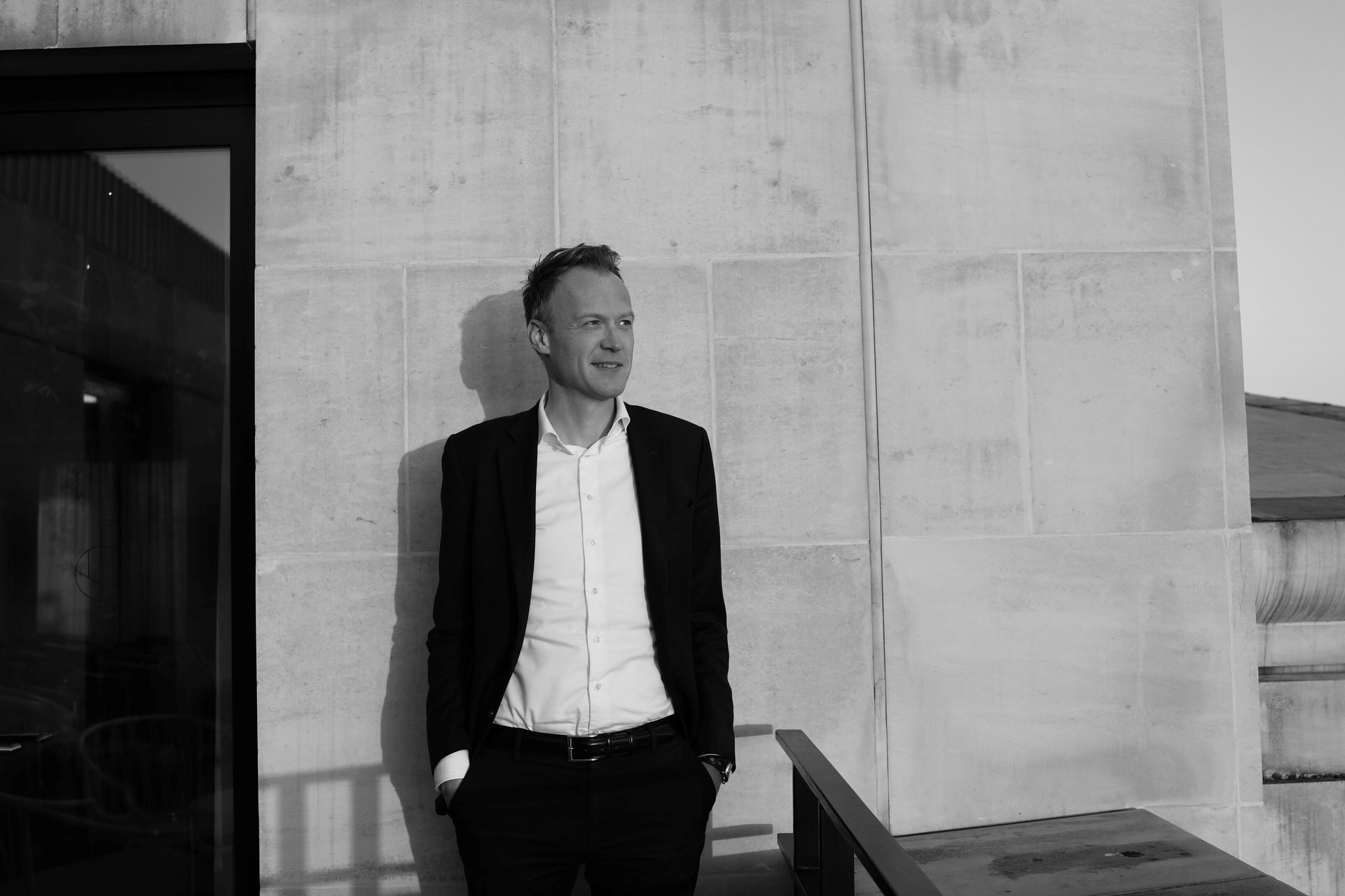 Black and white image of a man in a suit standing against a stone wall, looking to the right.
