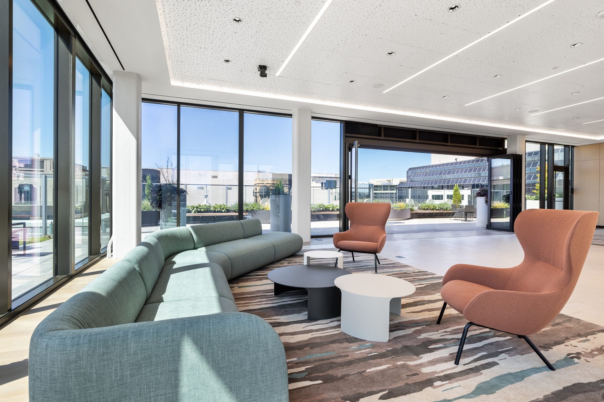 Modern lounge with light blue curved sofa, two orange armchairs, coffee tables, and large windows.