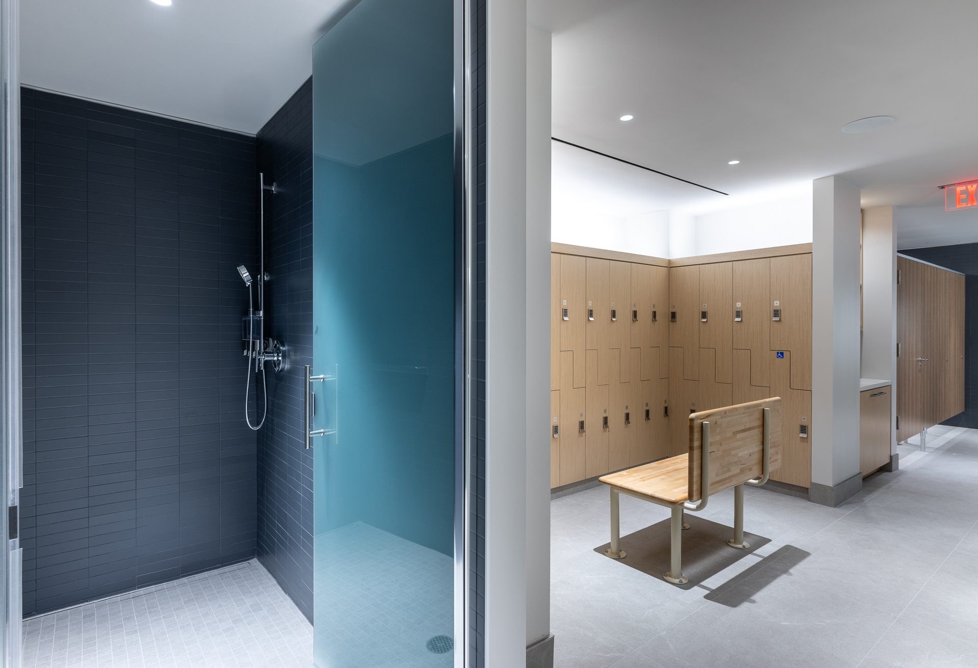 Modern changing room with a dark-tiled shower stall and wooden lockers, featuring a bench in the center.