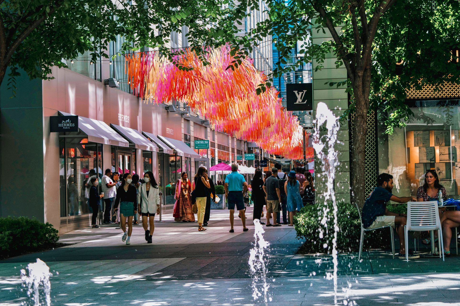 A lively outdoor shopping street with people, colorful ribbons overhead, luxury stores, and fountains.