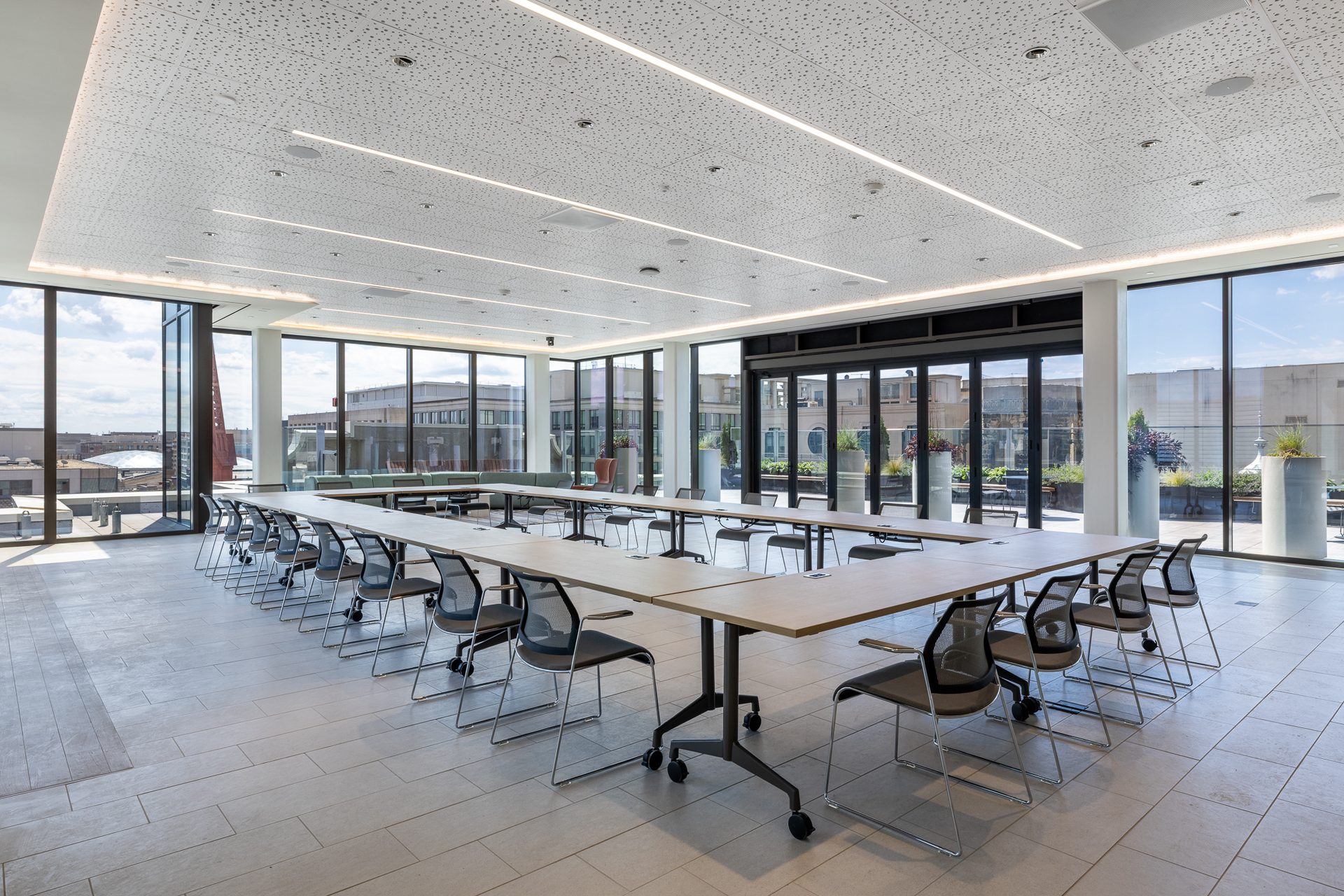 A spacious, modern conference room with U-shaped tables, mesh chairs, and large windows overlooking a city.