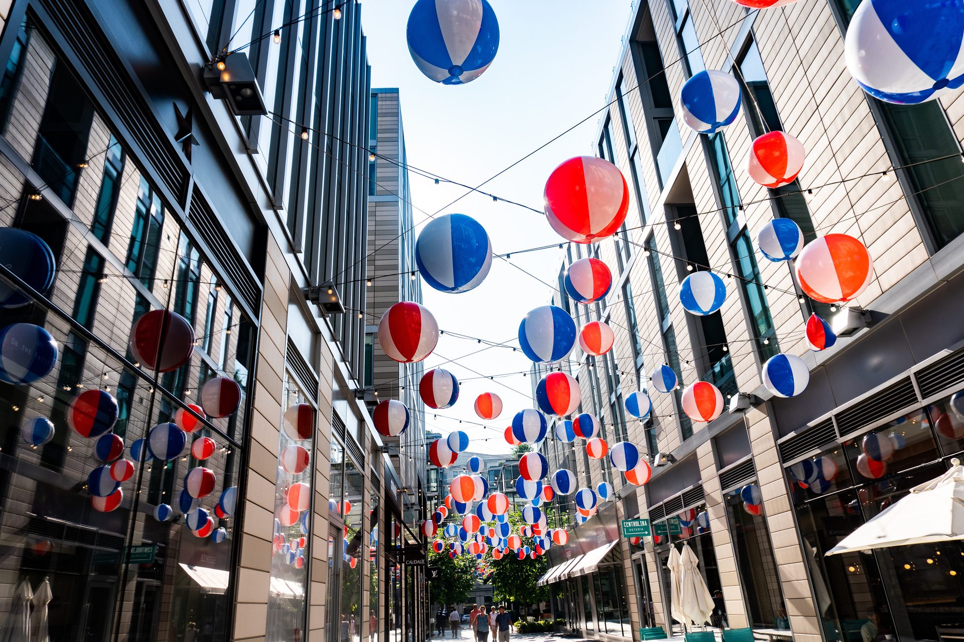 An outdoor pedestrian walkway in a city, decorated with strings of red, white, and blue beach balls overhead.