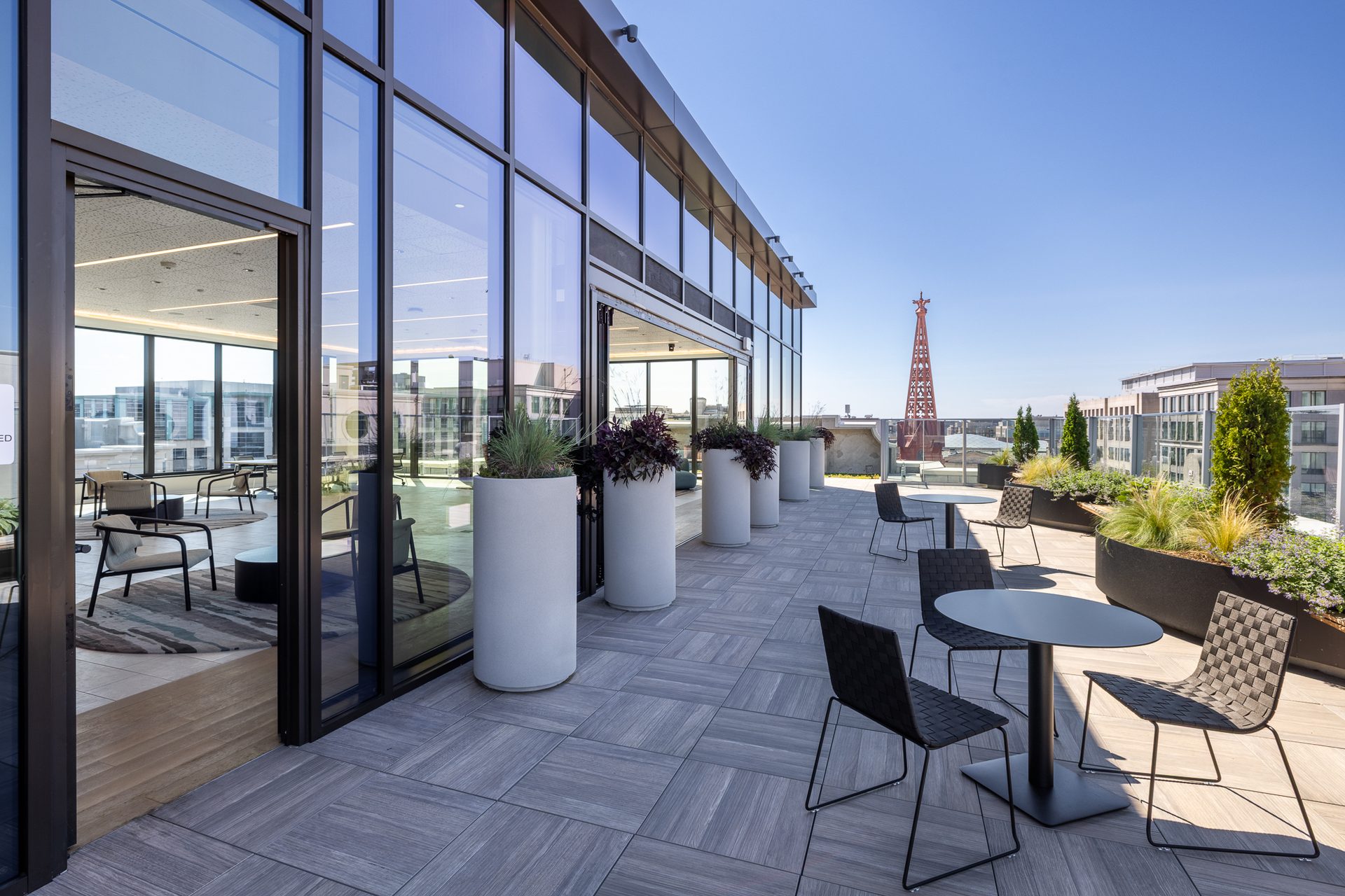 Modern rooftop terrace with outdoor seating, planters, floor-to-ceiling windows, and a city view with a red structure.