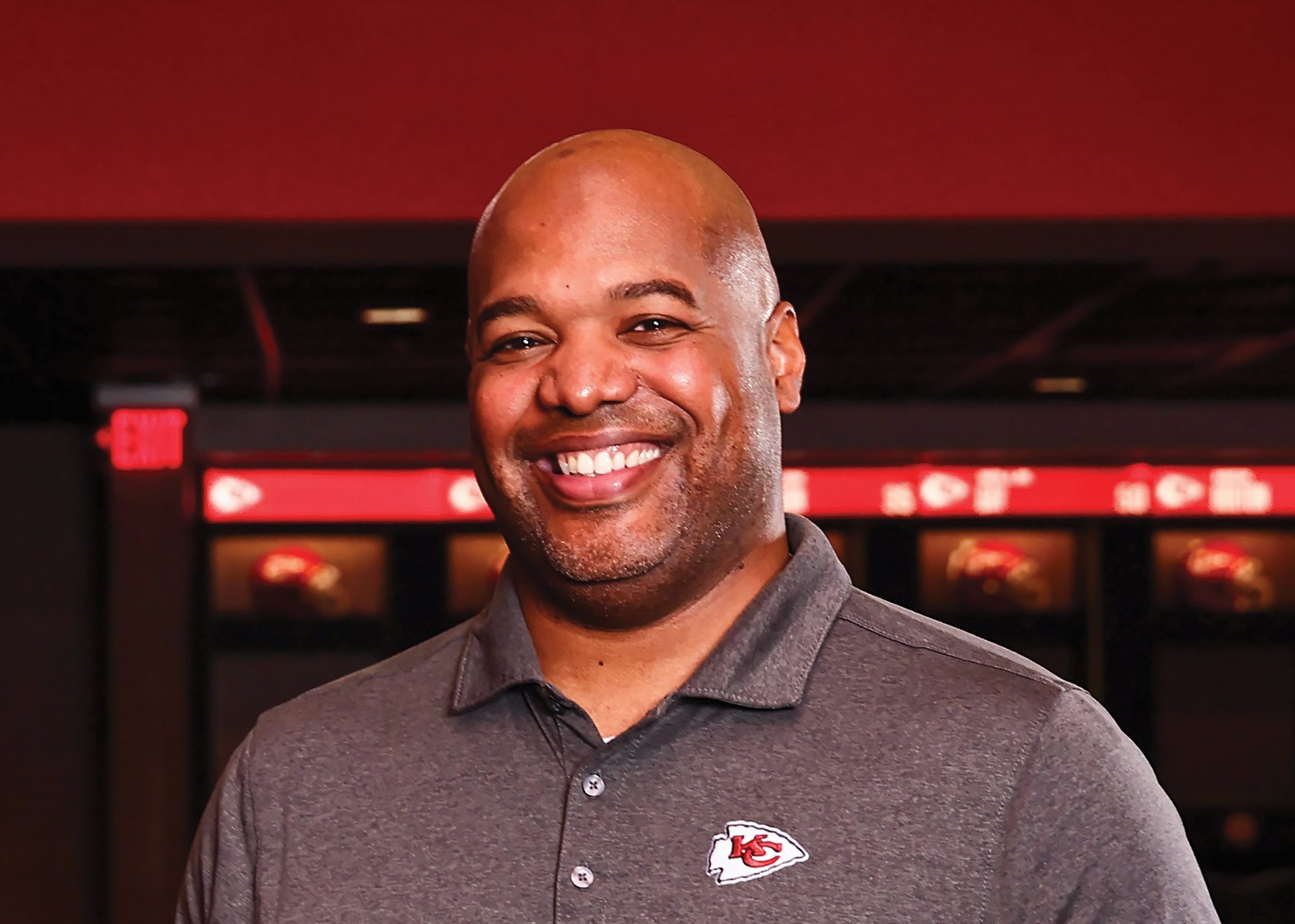 A bald black man smiles in an athletic facility, wearing a grey Kansas City Chiefs polo