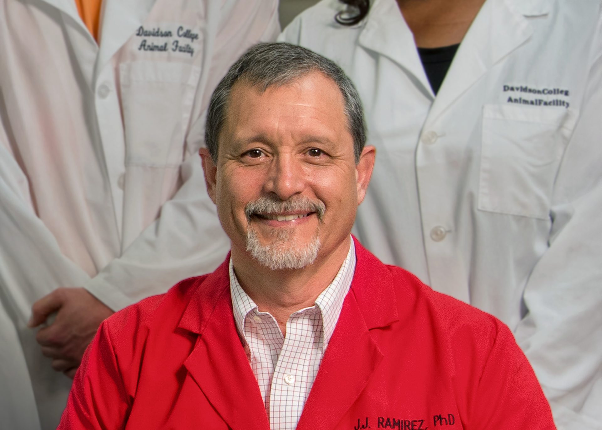 An older professor with a red lab coat, grey hair and facial hair, and tan skin sits in front of students