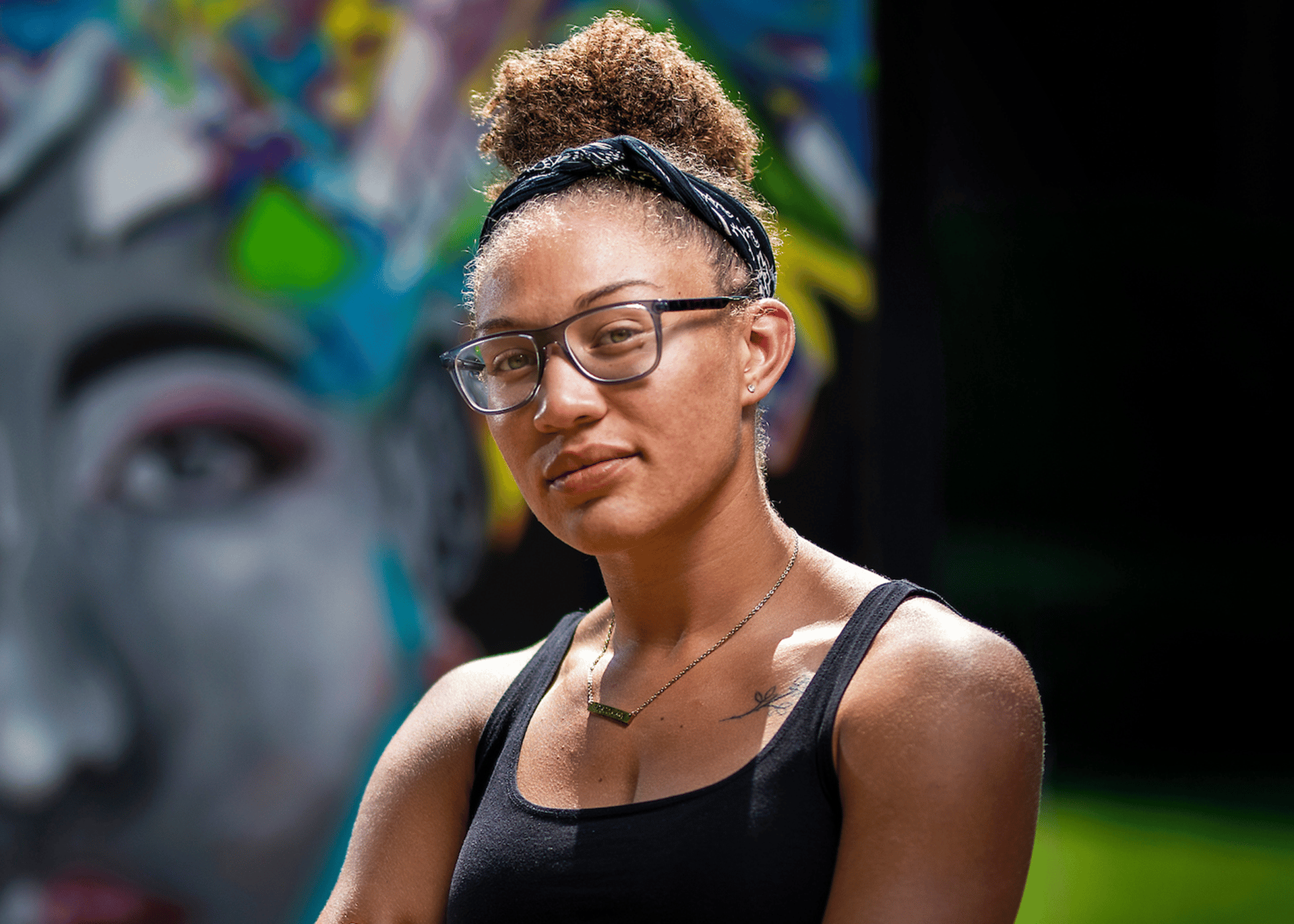 A young female artist with curly brown hair and medium skin tone stands in front of a mural.