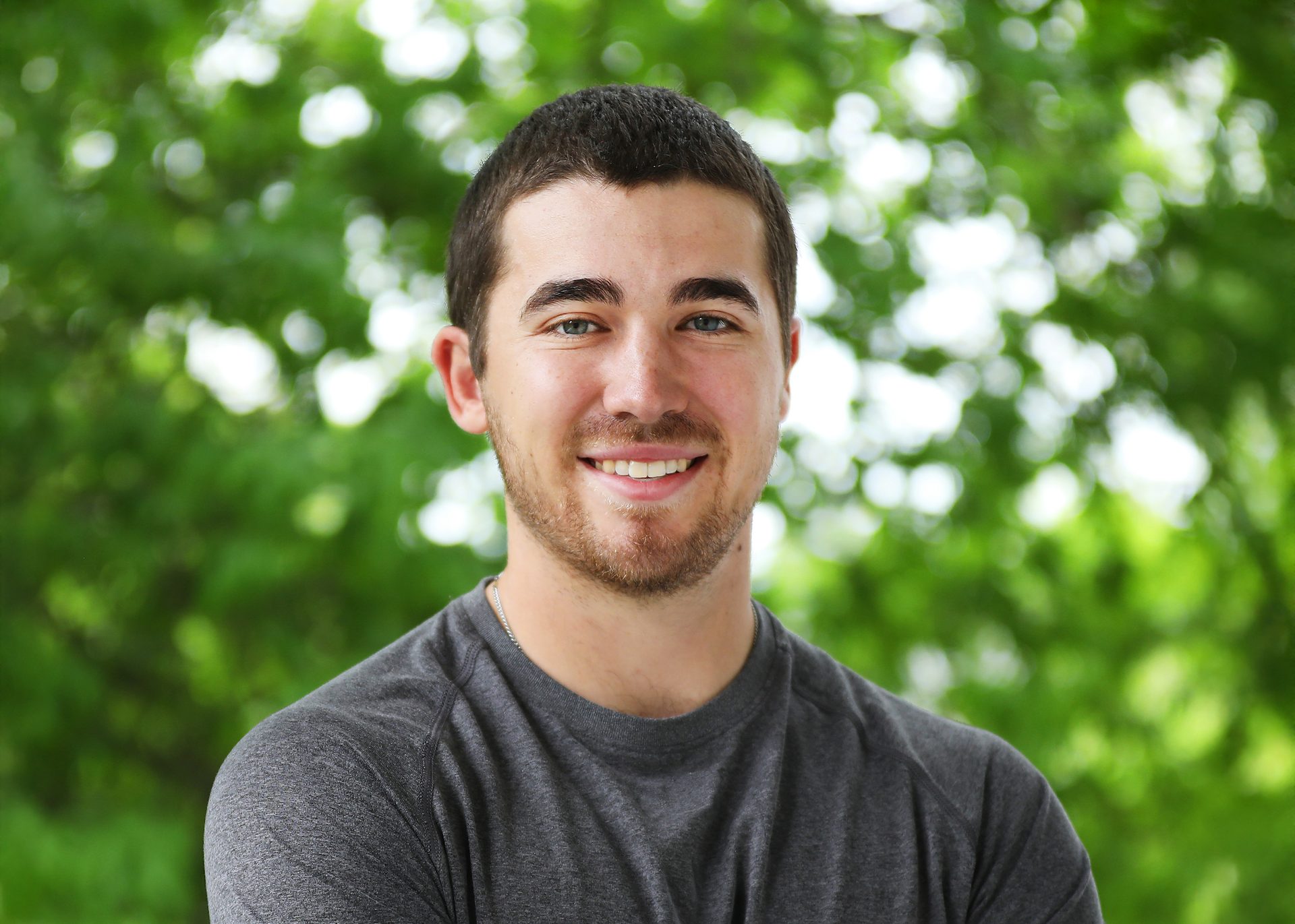 Smiling white male alumus, between 20-30 years of age, wearing grey t-shirt with brown hair