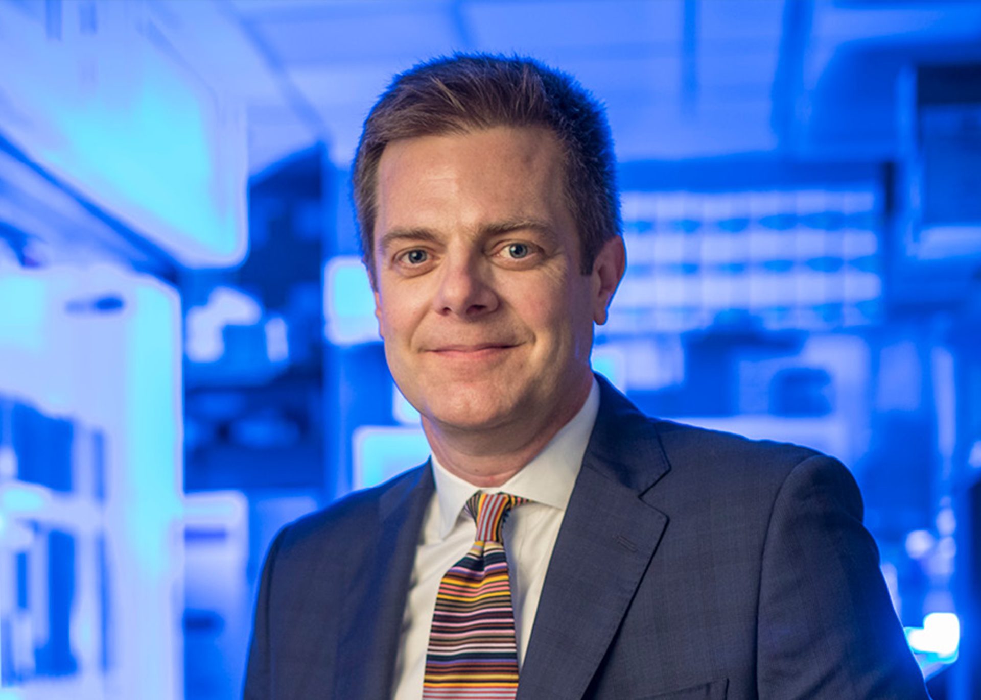 White man age range 25-45 with brown hair, a navy suit, and striped tie stands in a blue laboratory