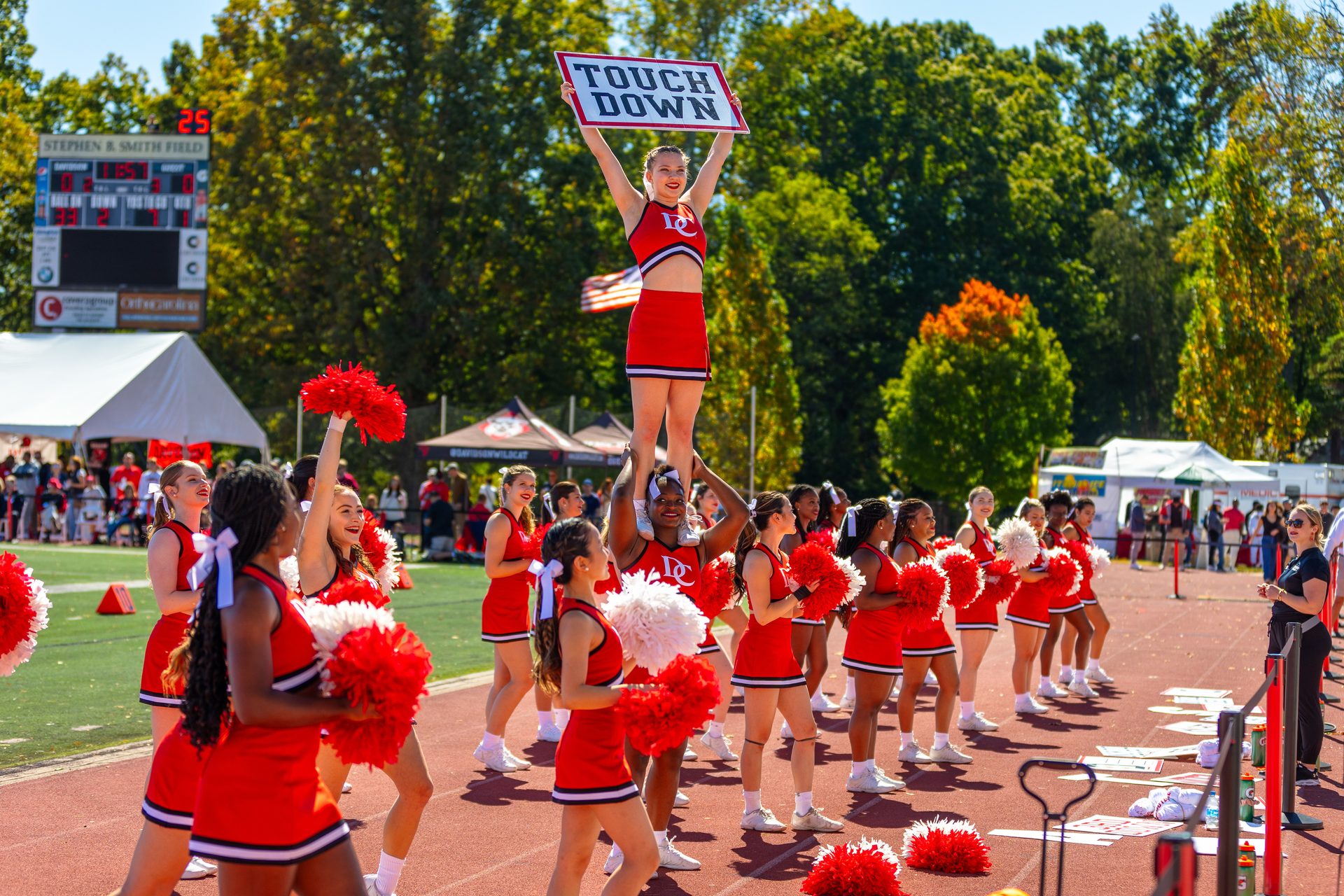 Cheerleaders perform on a sunny field; one holds a "TOUCH DOWN" sign while lifted by another.