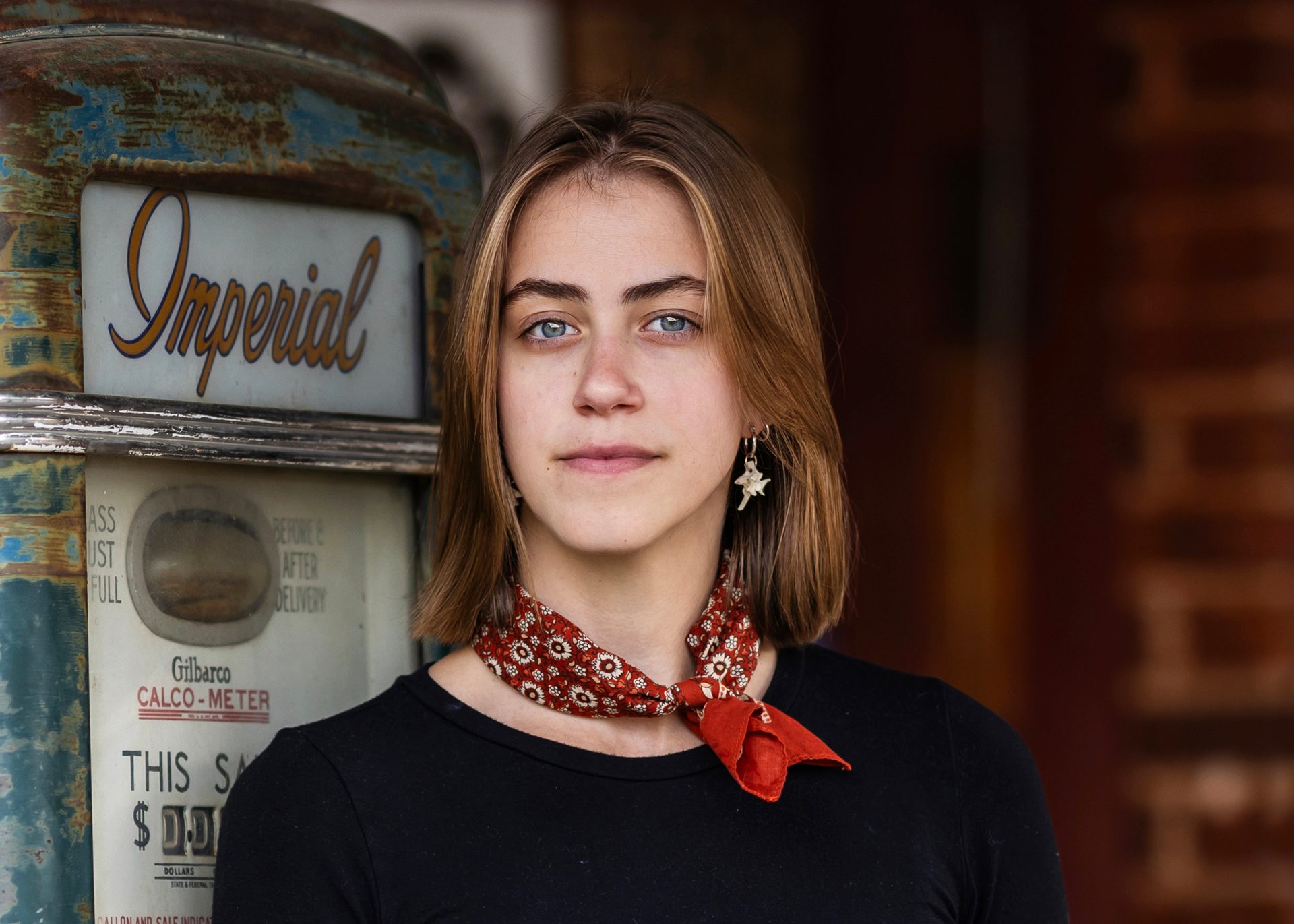 Recent graduate, female, with red bandanna and blonde, shoulder length hair poses in front of an antique gas pump
