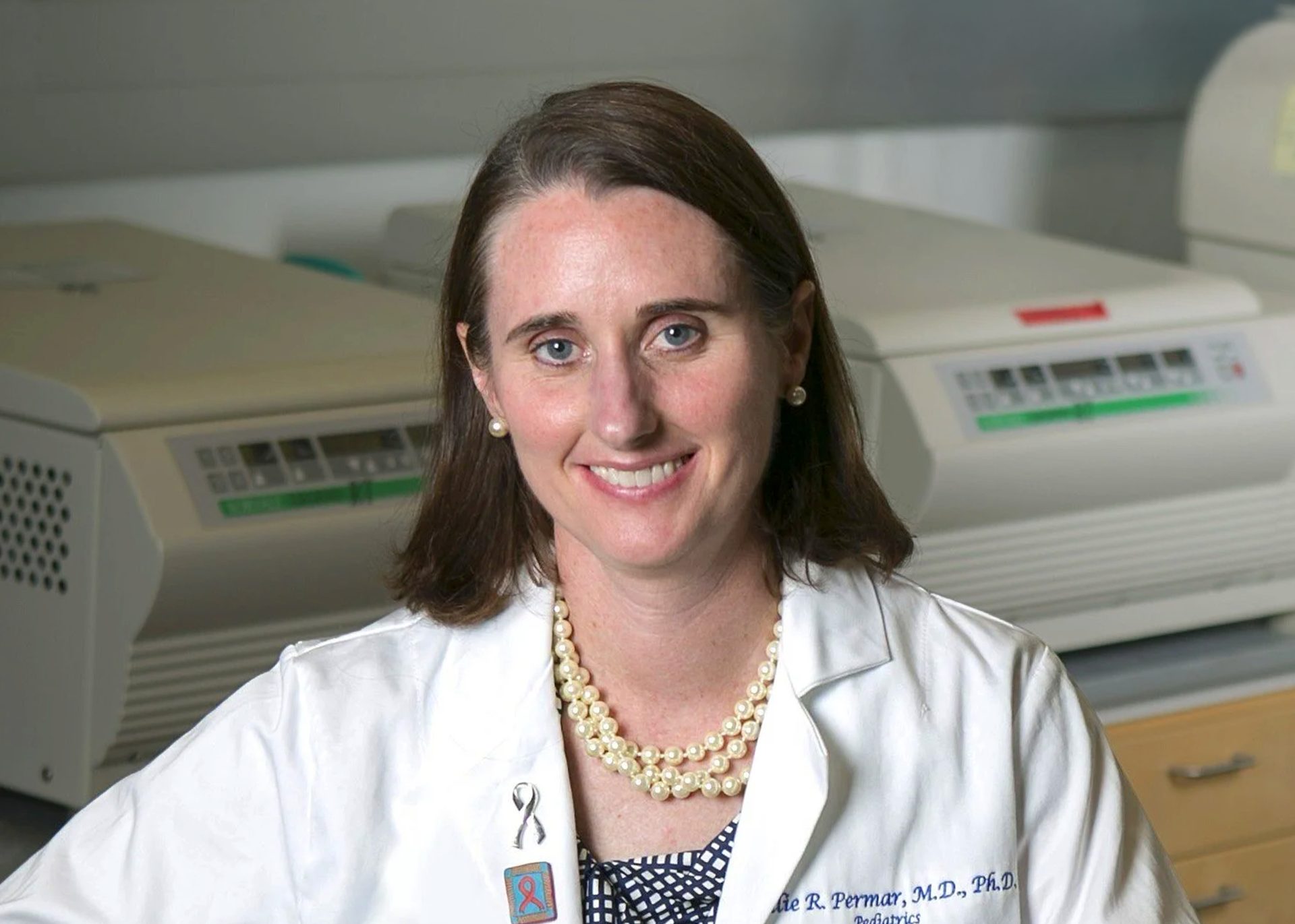 Female scientist with brown hair, approximately 29-40 years of age, wearing a white lab coat and smiling