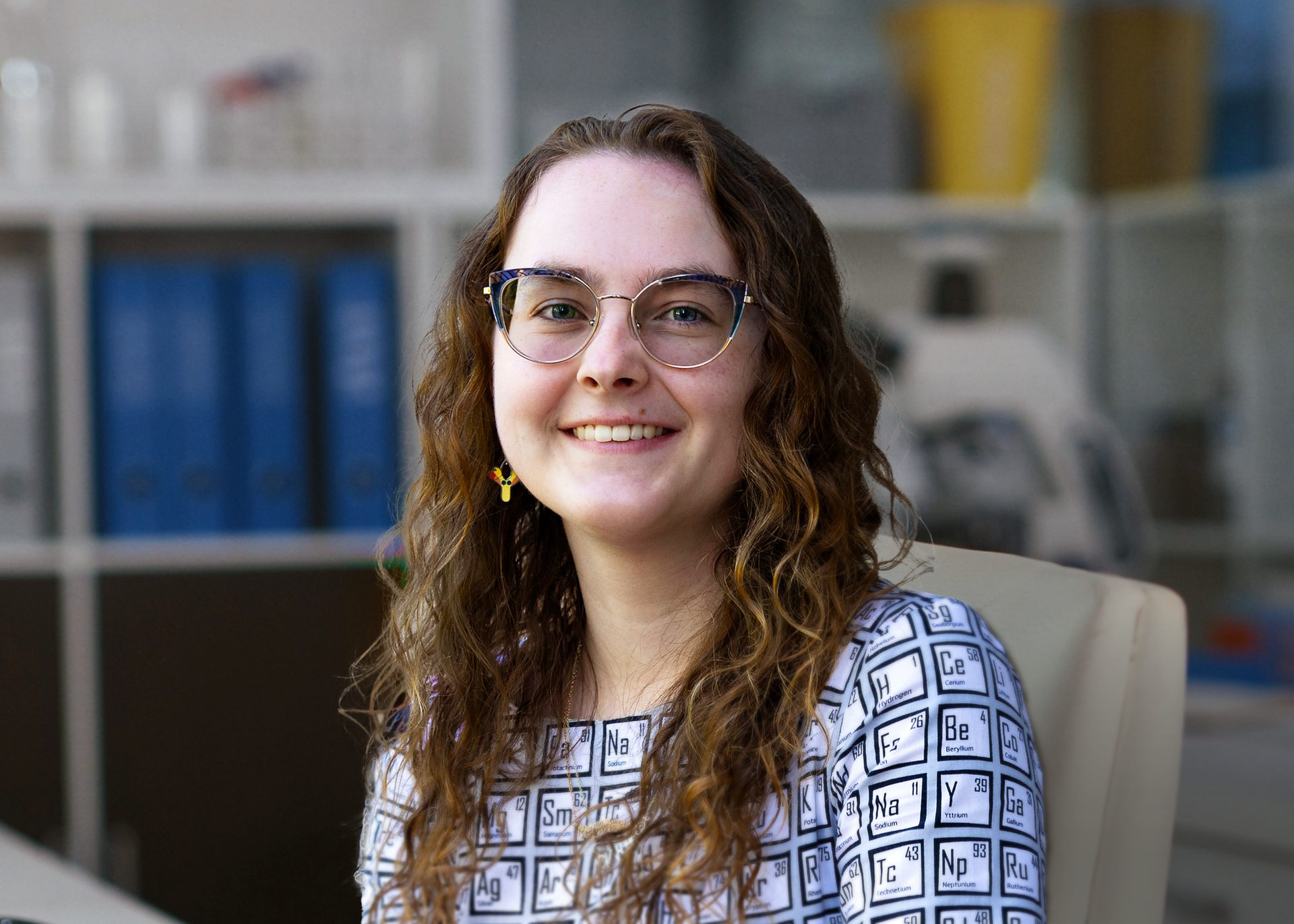 Female student with glasses, long curly light brown hair wears a pattern resembling the periodic table of elements