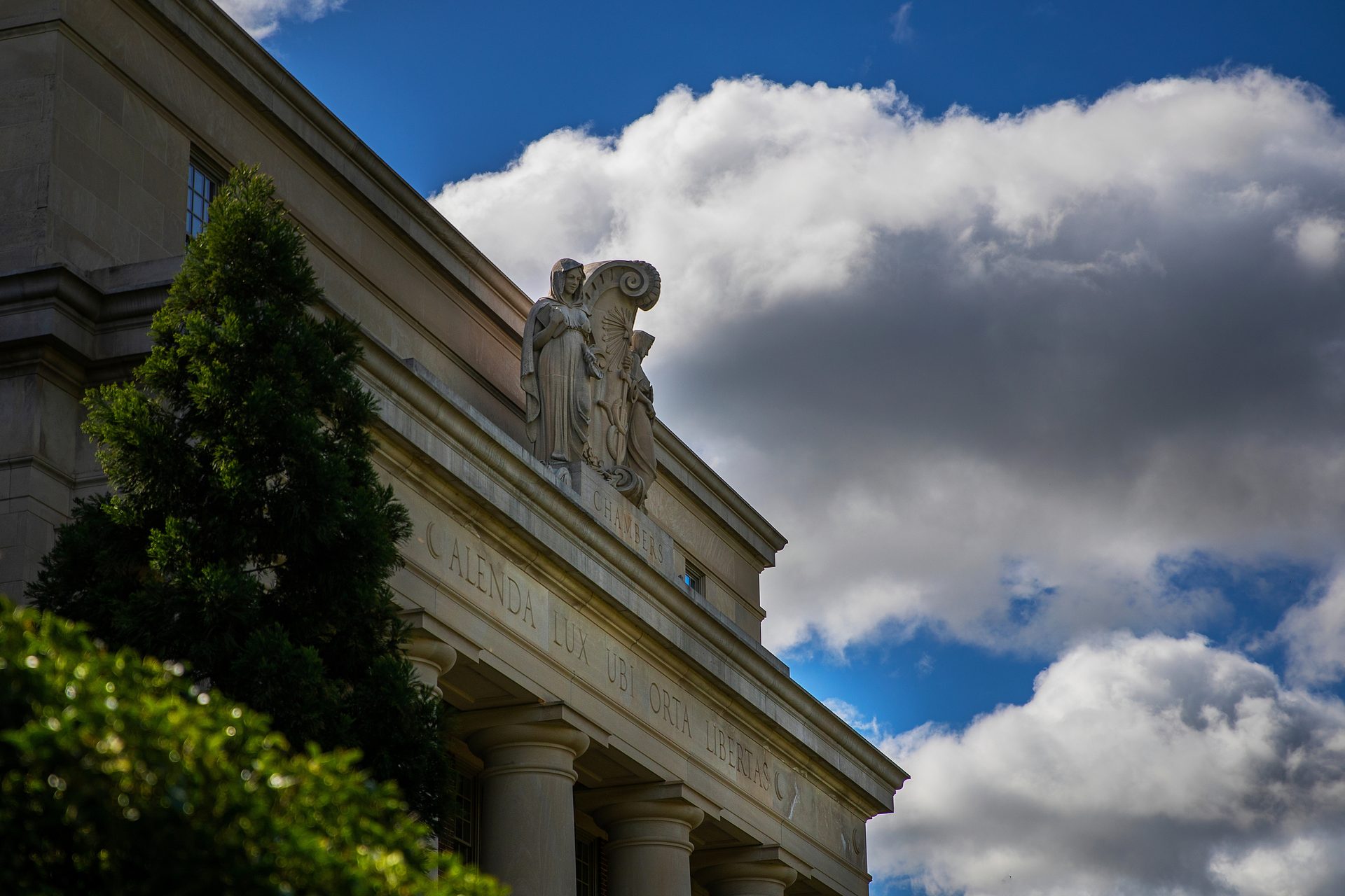 Classical stone building with statues and Latin inscription under cloudy sky.