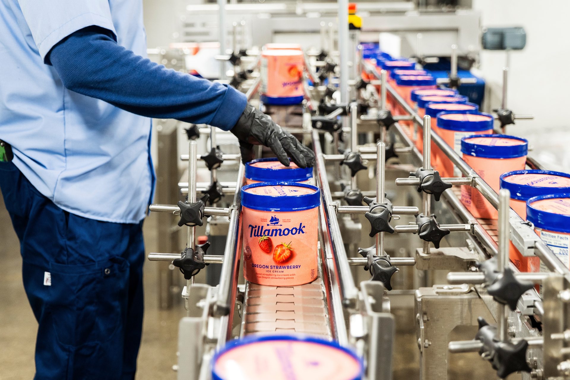 Worker guiding Tillamook Oregon Strawberry ice cream tubs on a factory conveyor belt.