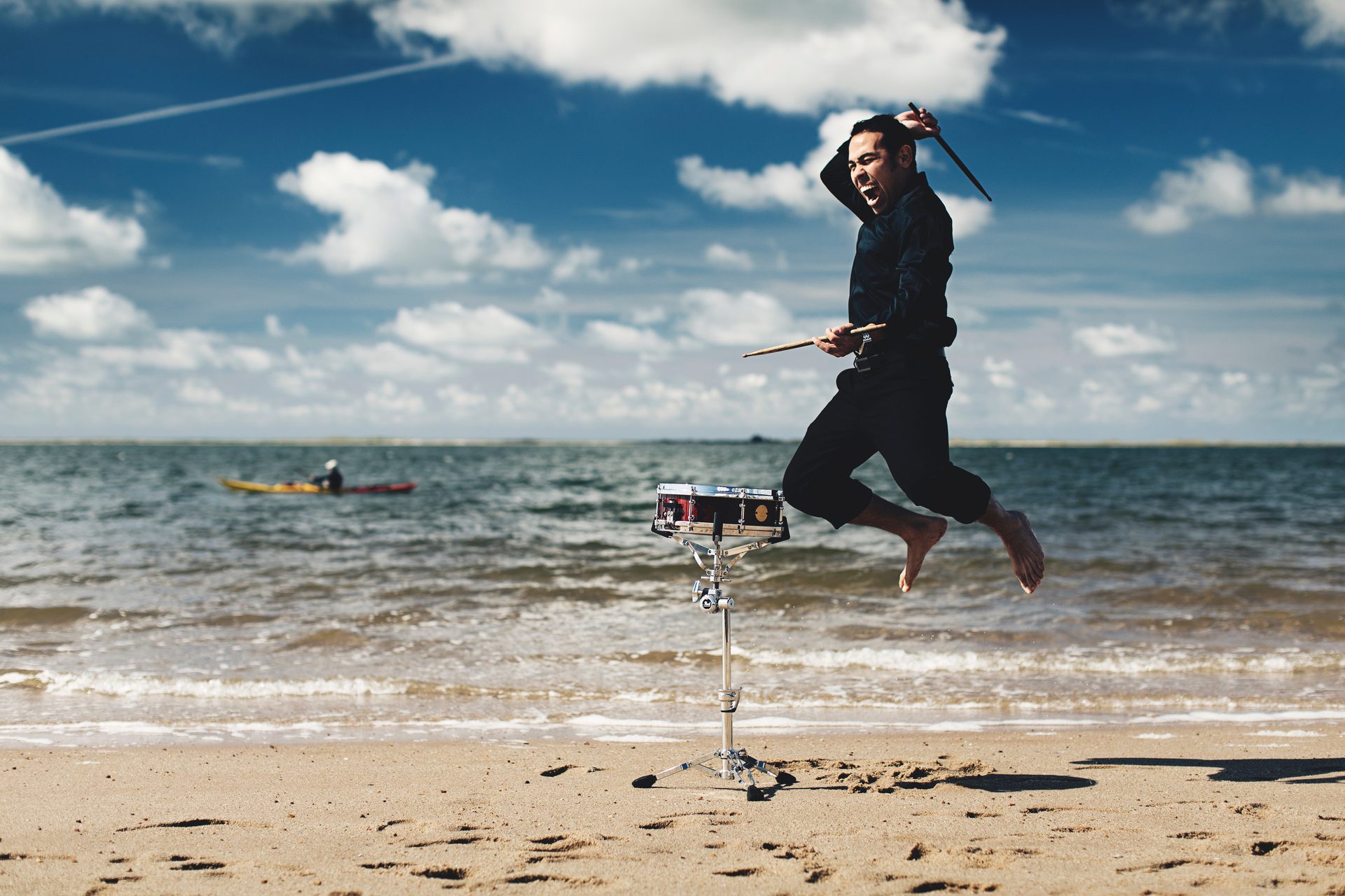 Man jumping mid-air while playing a snare drum on a beach with the sea in the background.