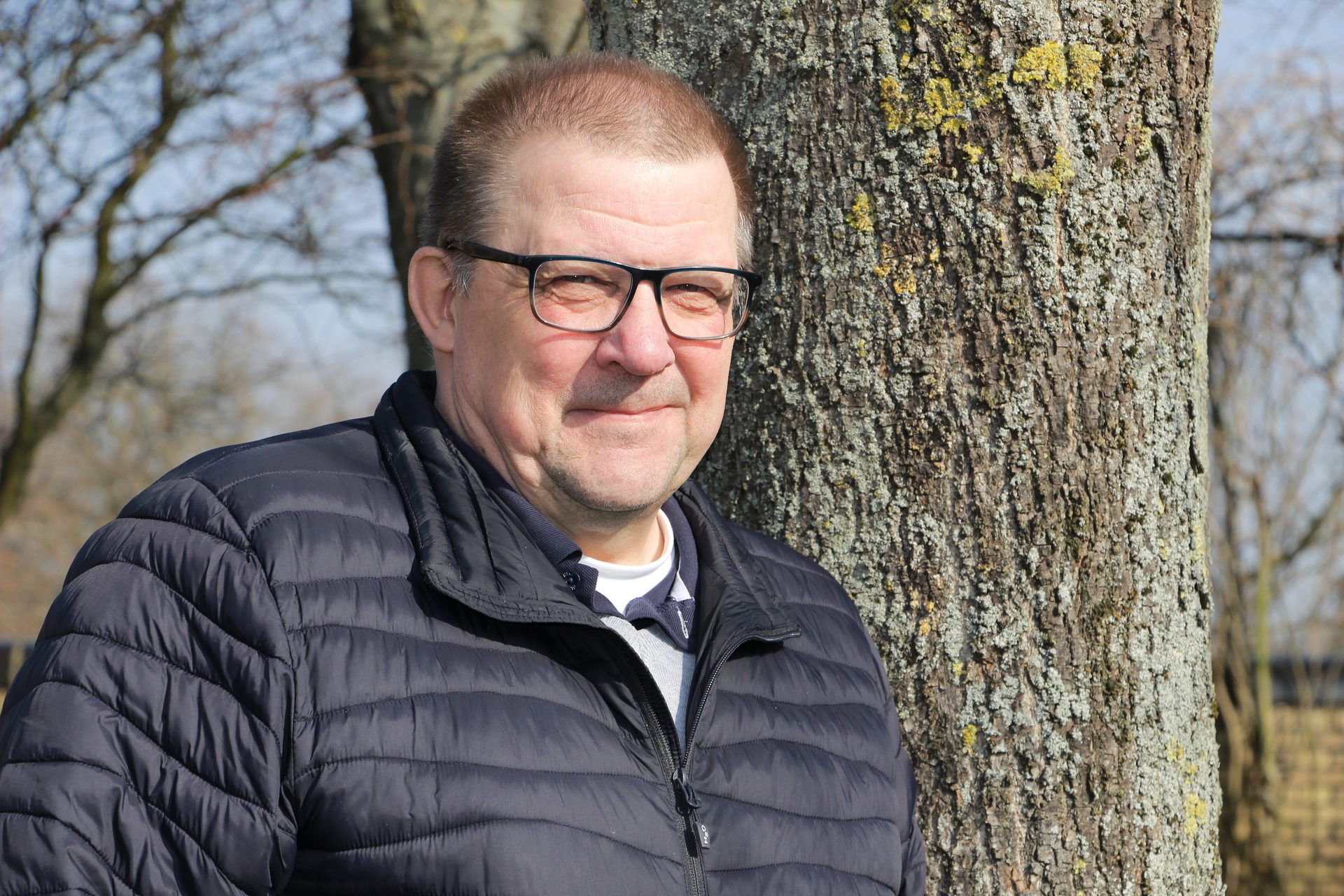 An older man with glasses and a puffer jacket smiles by a tree on a sunny day.