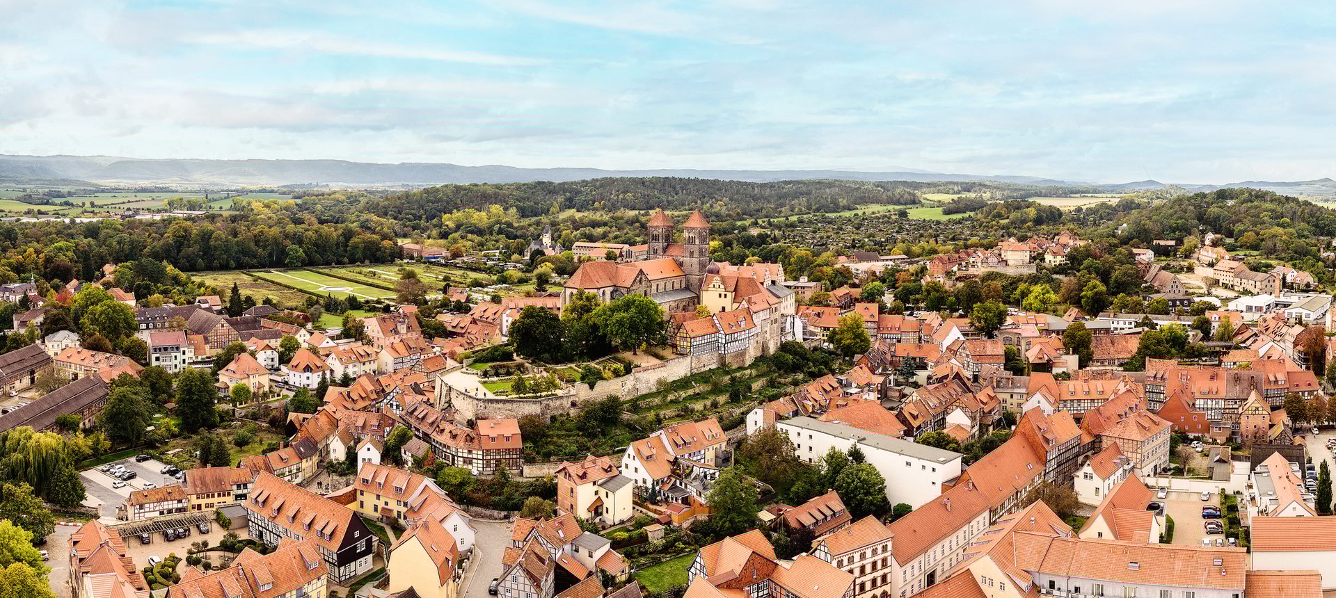 Aerial panorama: historic European town with red roofs, a large church, and green hills.