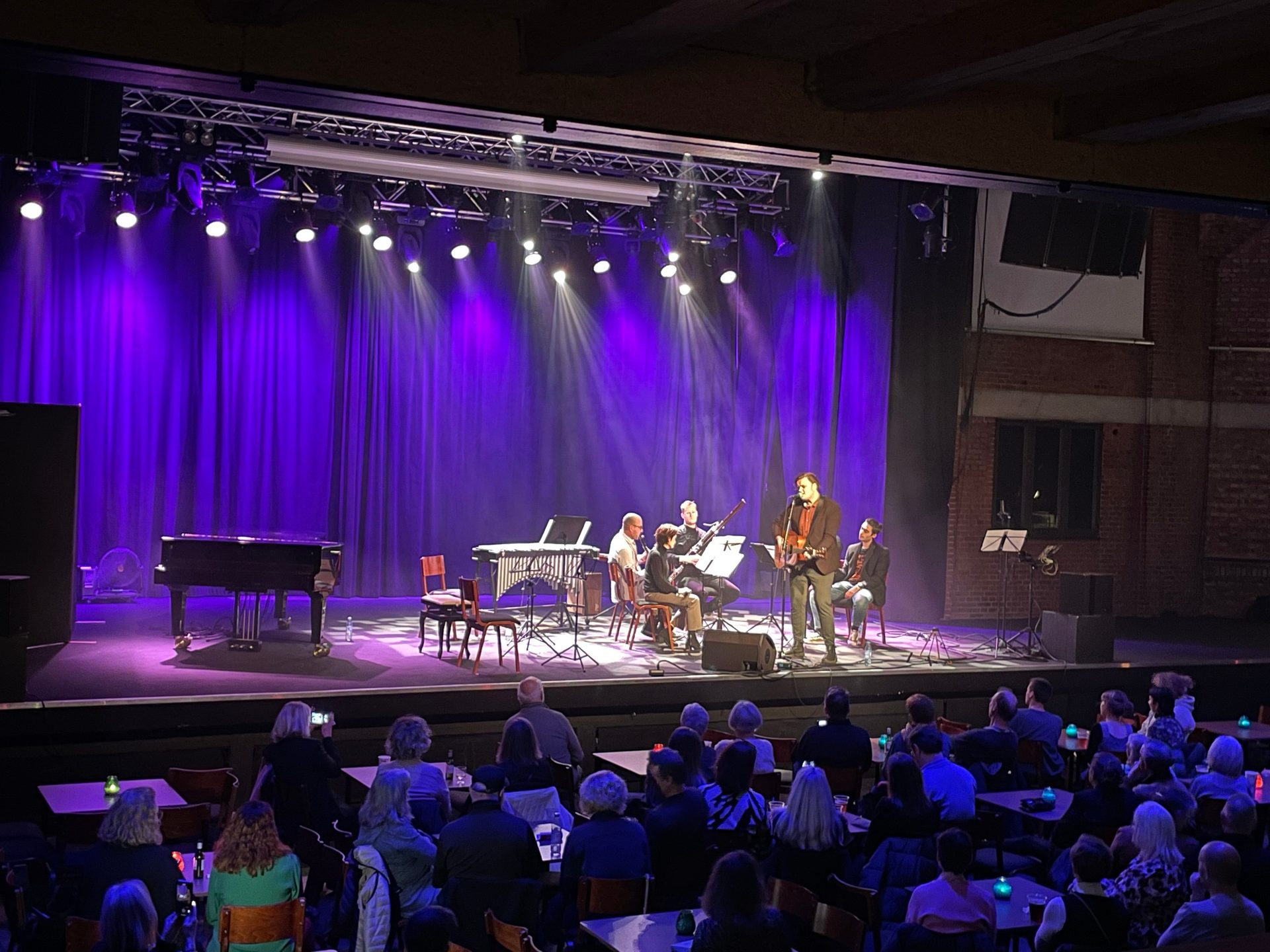 Musicians perform on a purple-lit stage to an audience, featuring a singer with a guitar.