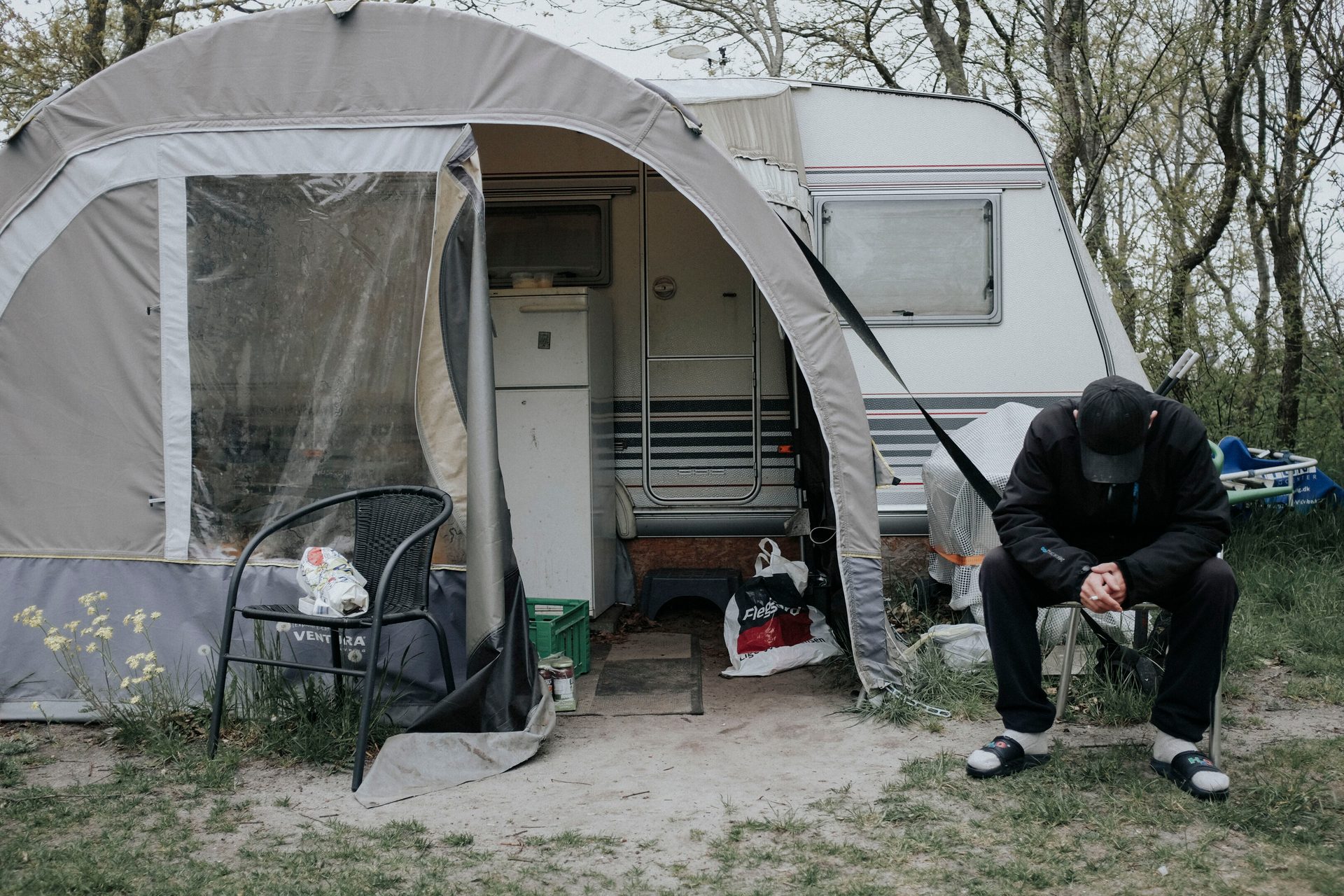 A person sits, head down, outside a camper van with an awning.