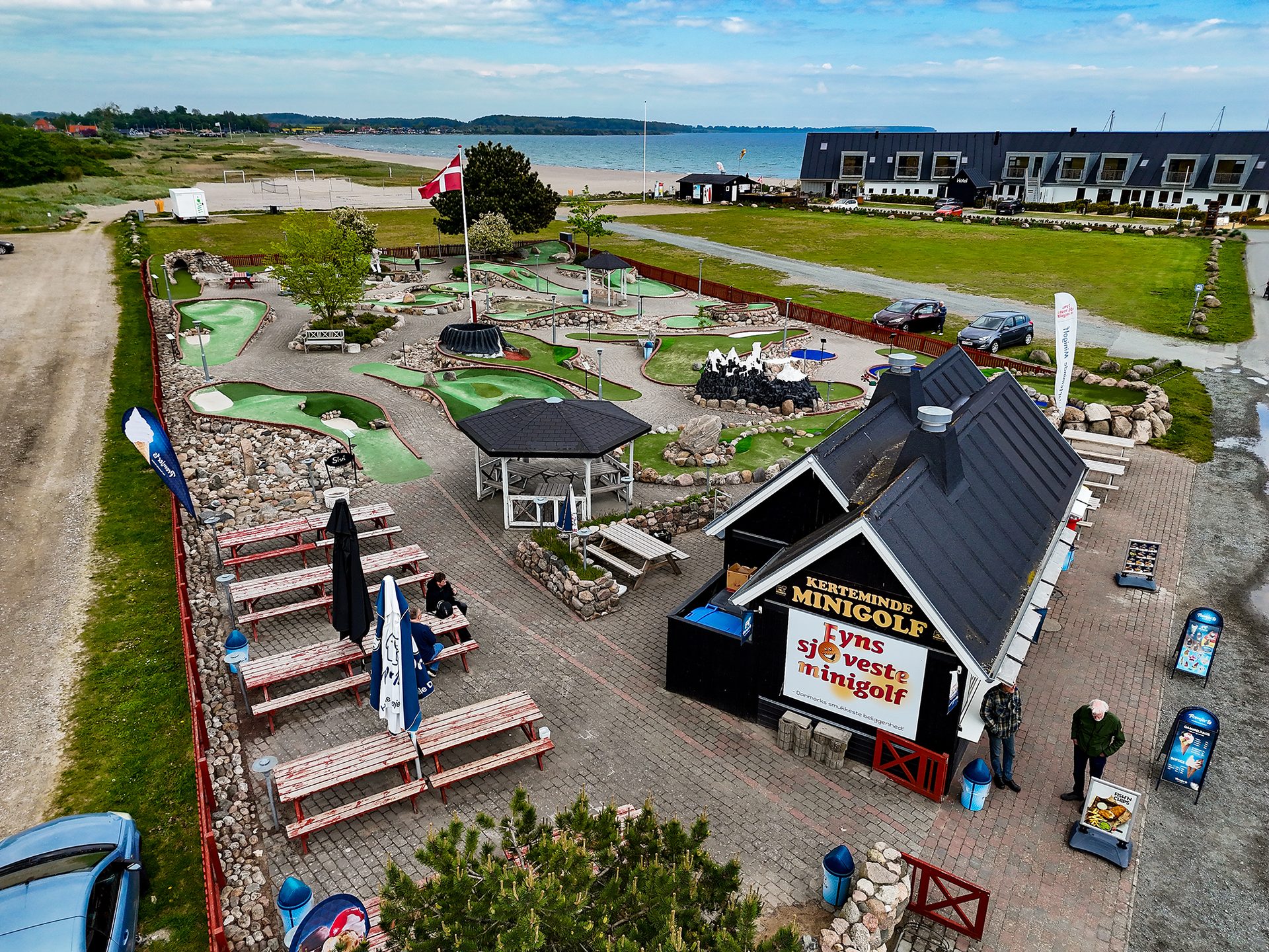 Aerial view of a colorful minigolf course, kiosk, beach, and hotel, with people playing and relaxing.