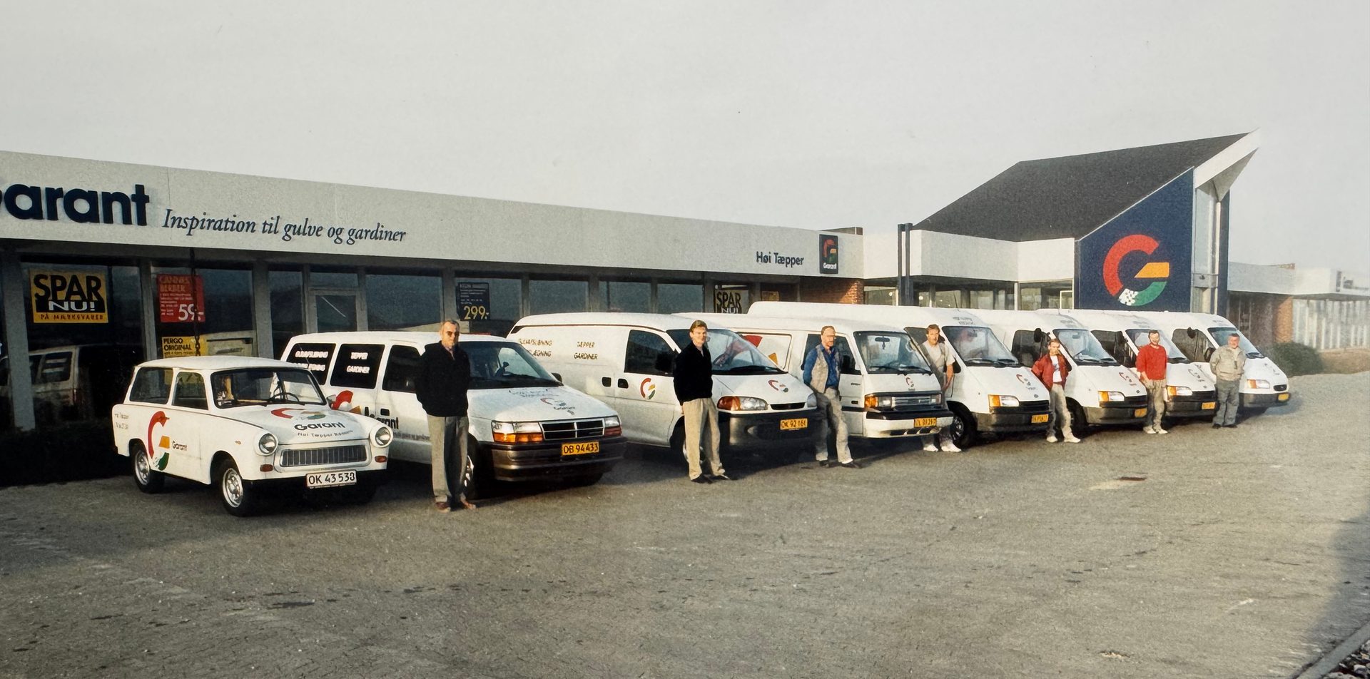 A group of employees posing with their white company vehicles, including vans and a small car, in front of their store.