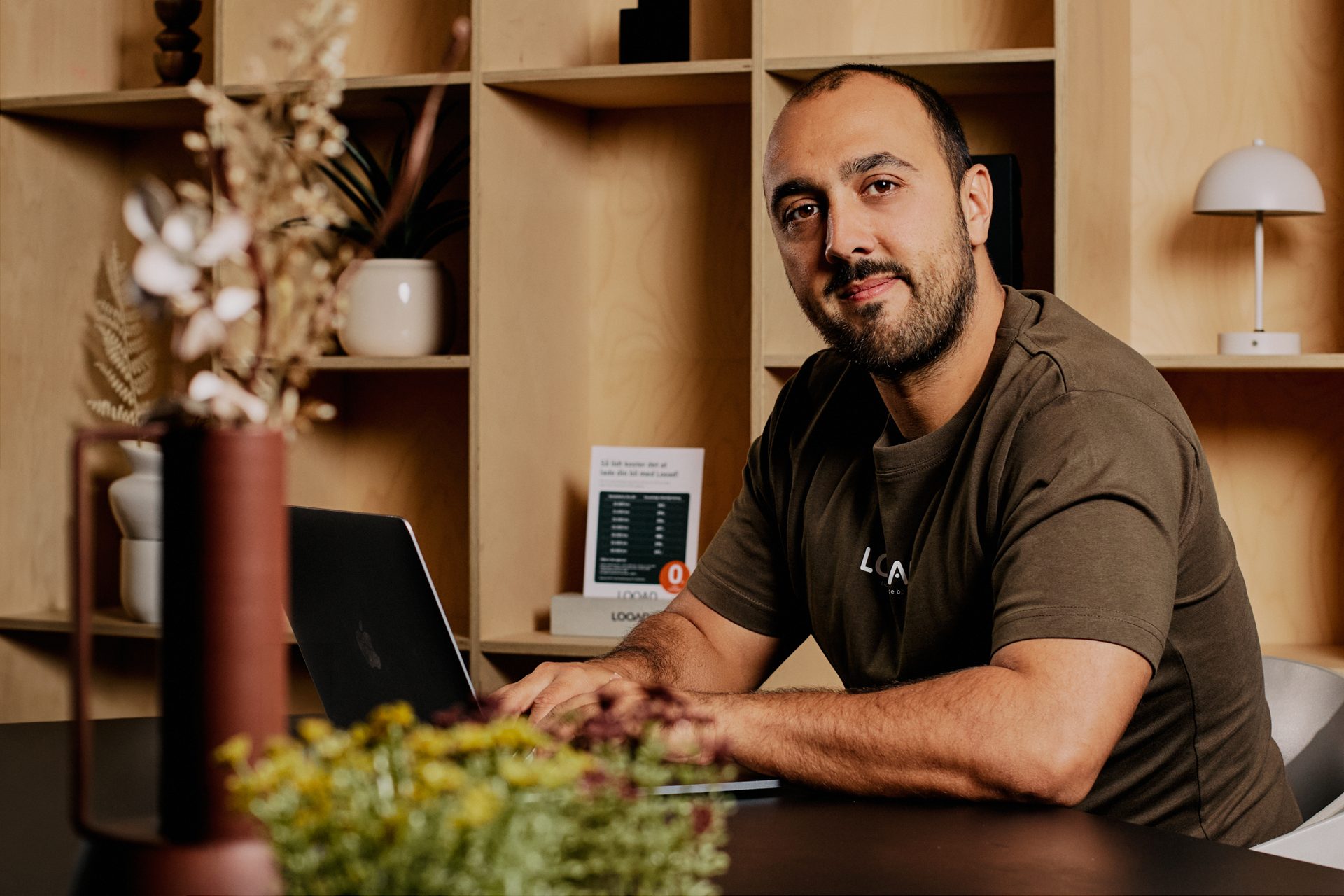 A man with a beard, wearing a dark green t-shirt, works on a laptop at a desk, looking at the camera.