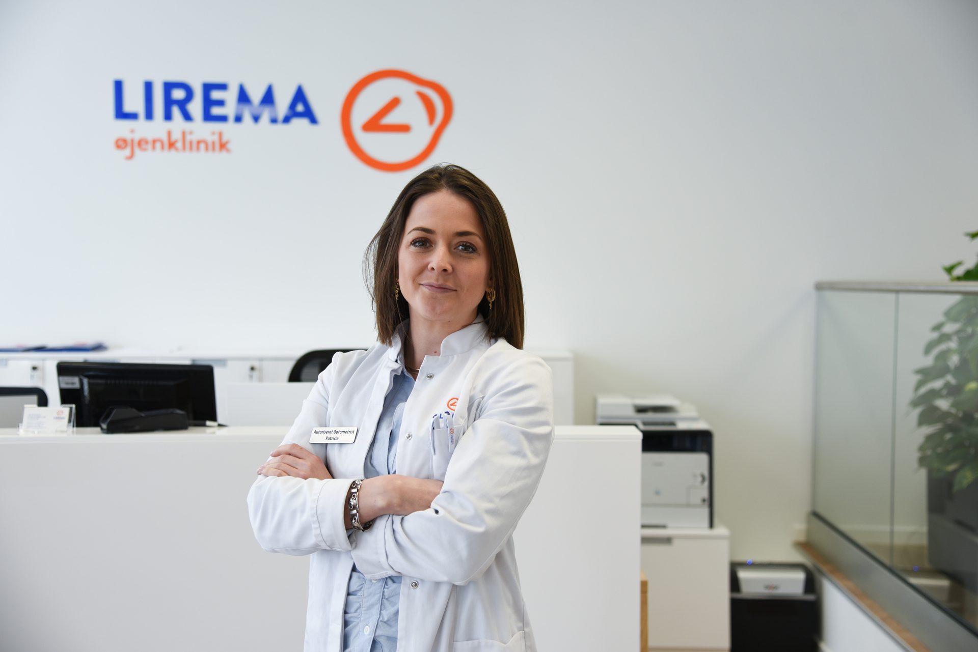 A woman in a white lab coat smiles with crossed arms in a modern clinic with "LIREMA øjenklinik" logo.