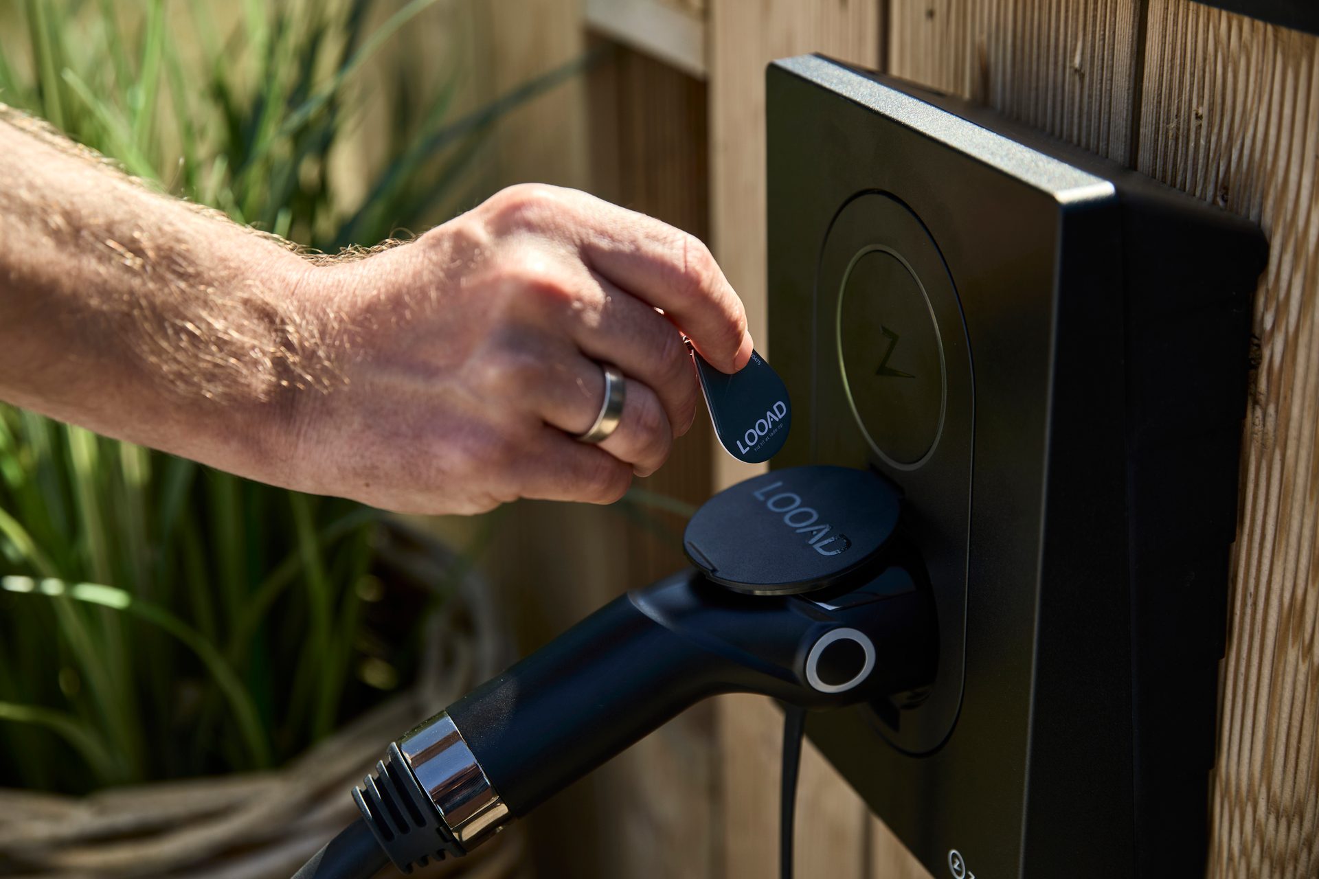 A hand activates an EV charger on a wooden fence using a small LOOAD fob.