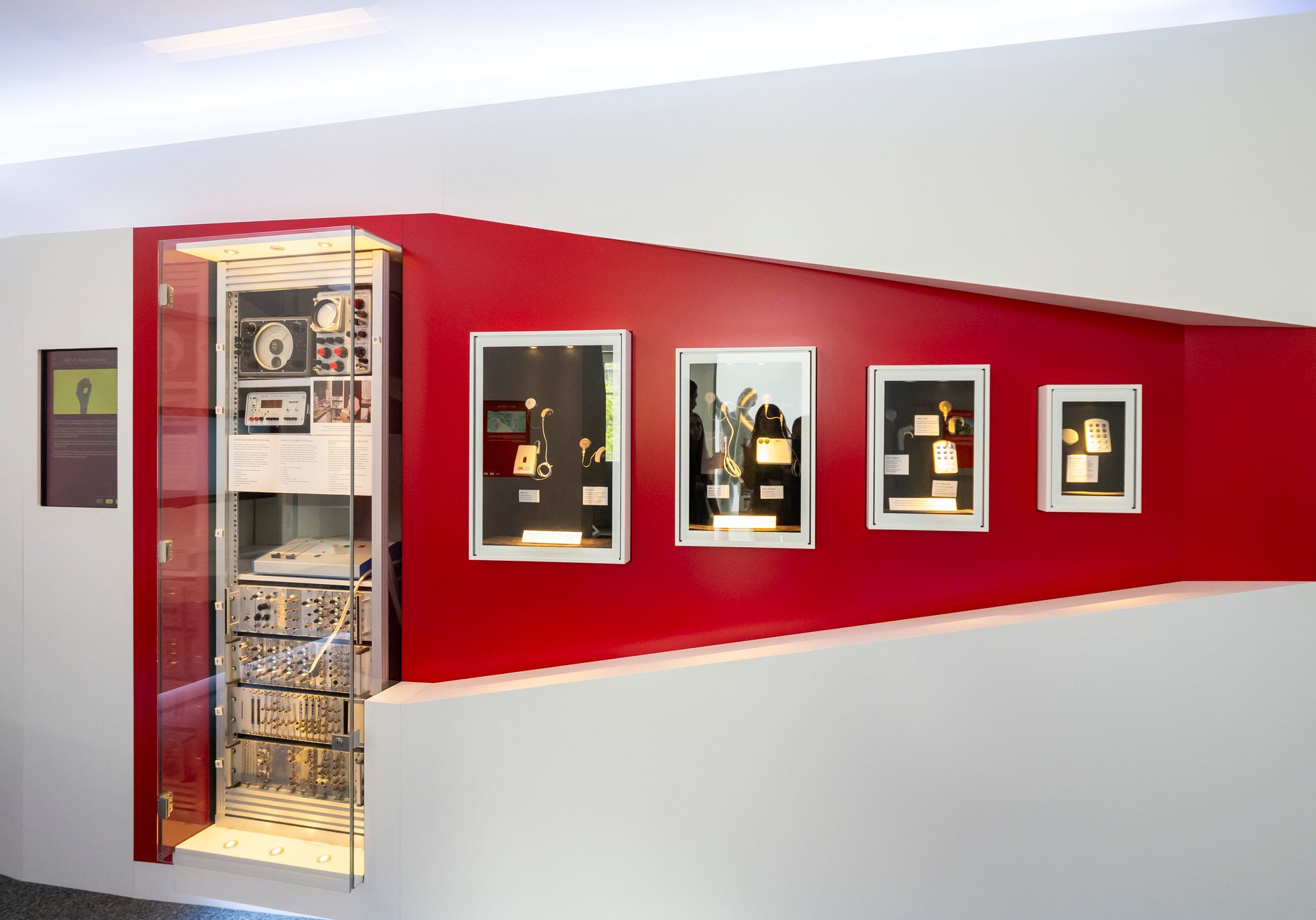 An exhibition displays vintage electronic equipment in a tall glass cabinet and smaller framed cases on a red wall.