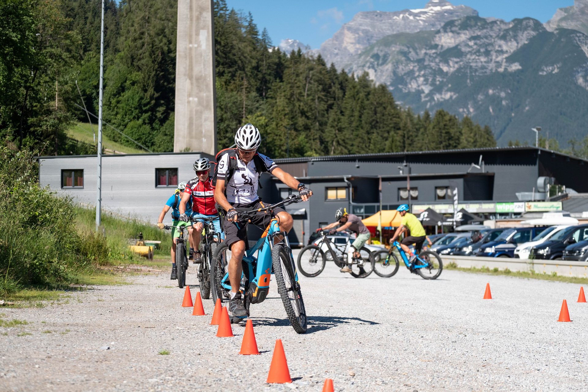Group of mountain bikers riding on a gravel path with orange cones, mountains in background.