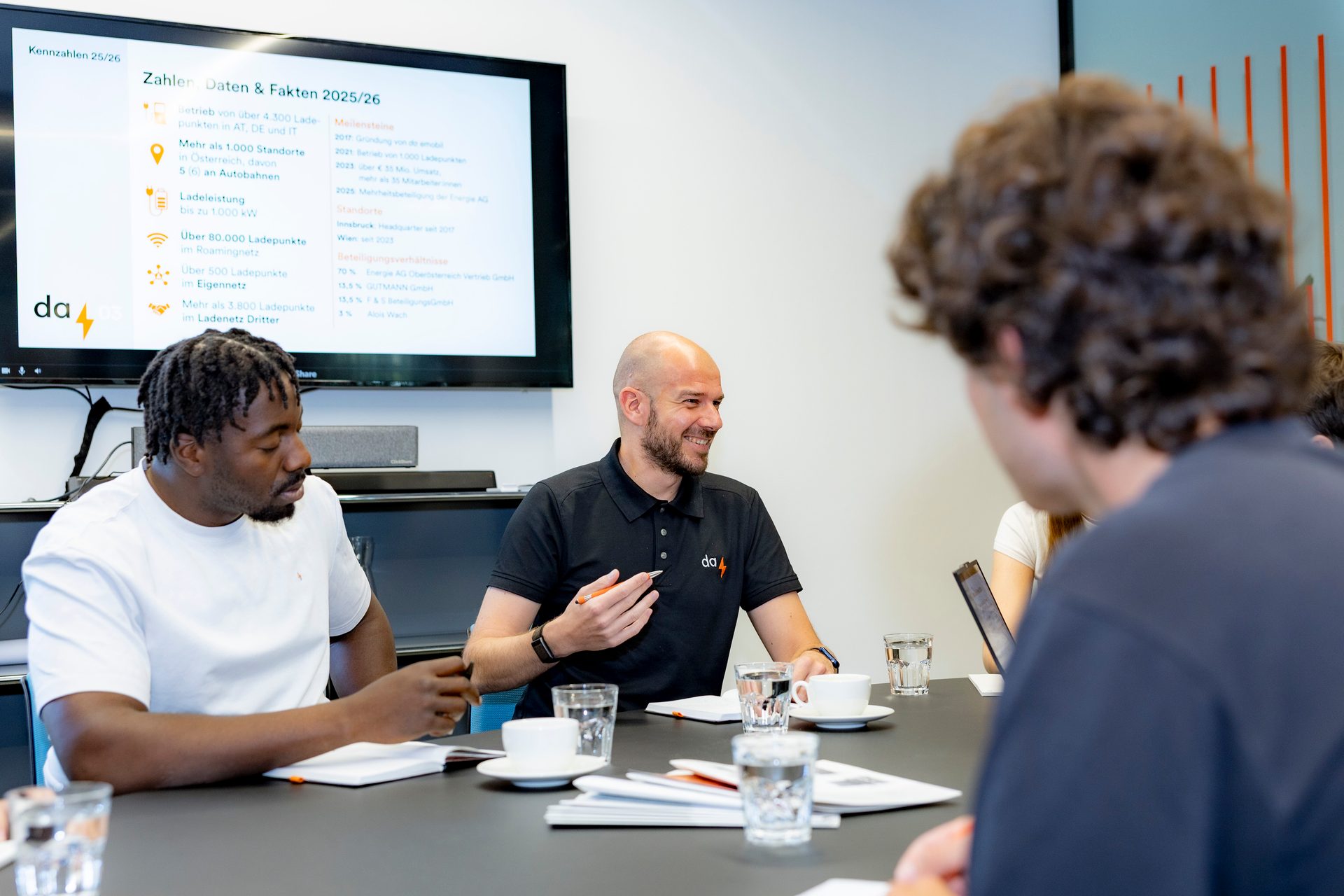 Three men in a bright office discussing around a table, with a presentation screen in the background.