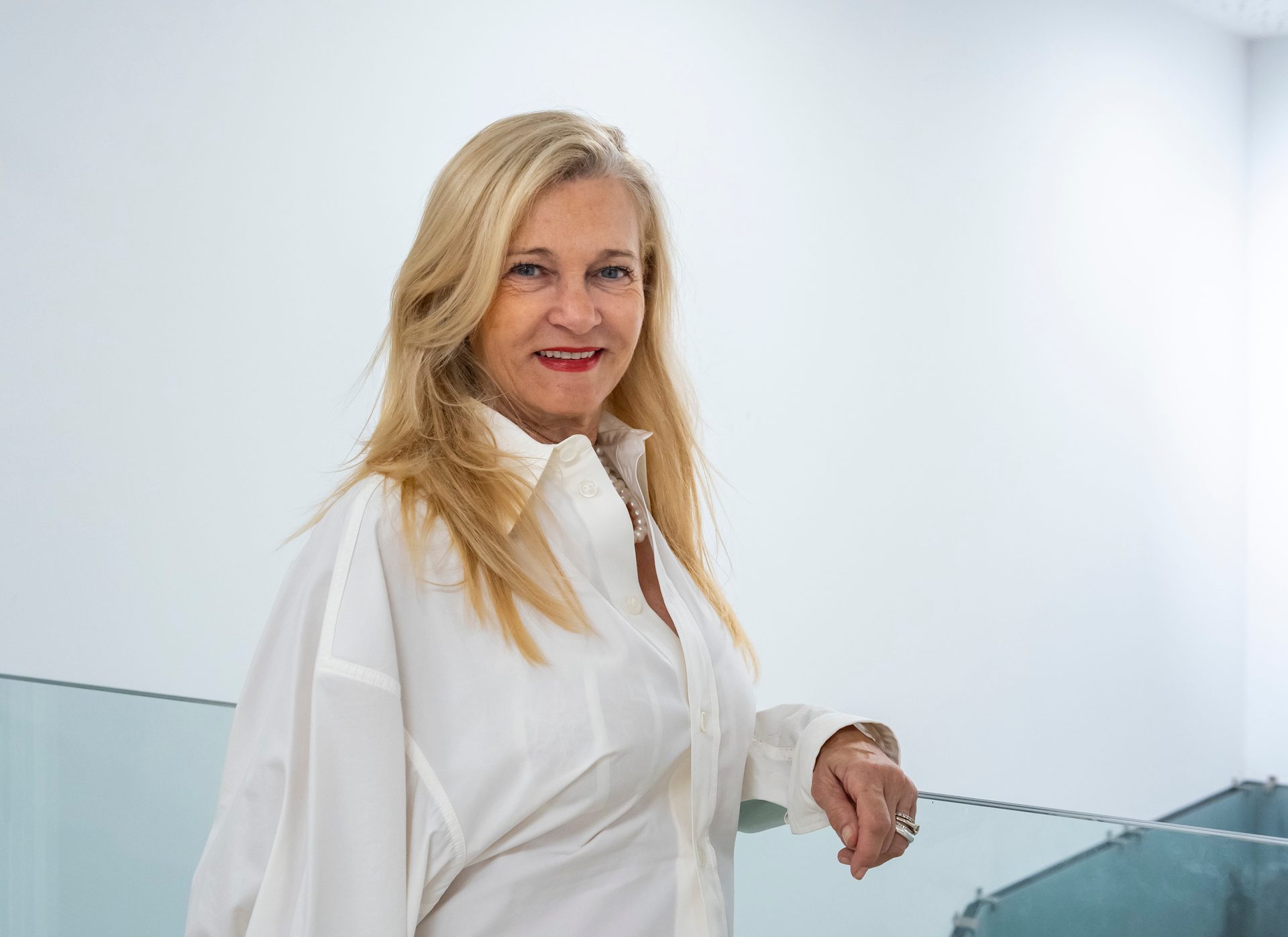 A smiling blonde woman in a white shirt and pearl necklace leans on a glass railing in a bright room.