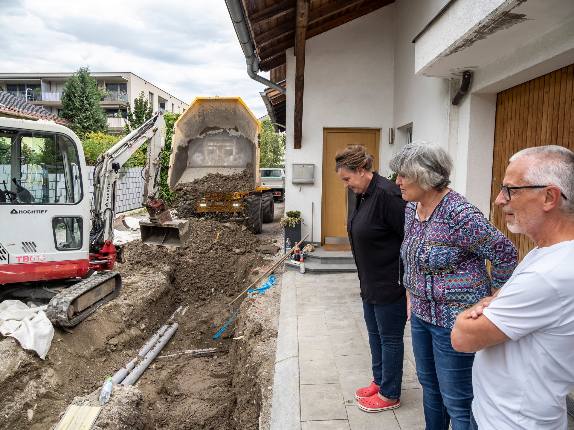 Three people observe an excavator and dump truck digging a trench near a house.