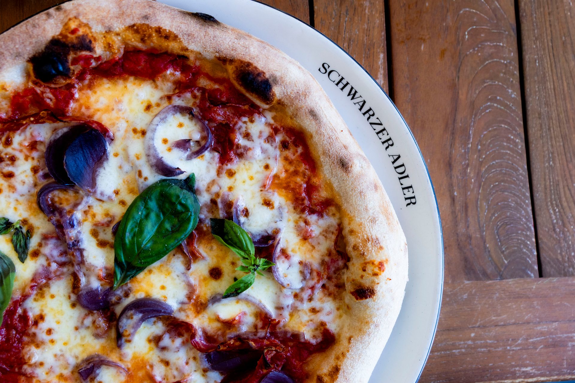 Delicious pizza with cheese, red onions, and basil on a plate with "SCHWARZER ADLER" text.