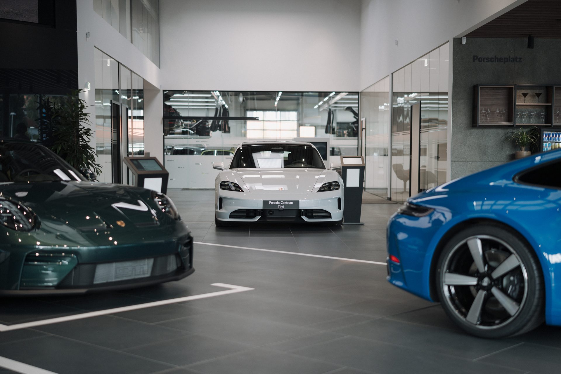 Three Porsche cars, including a white Taycan, green 911, and blue 911, displayed in a modern showroom.