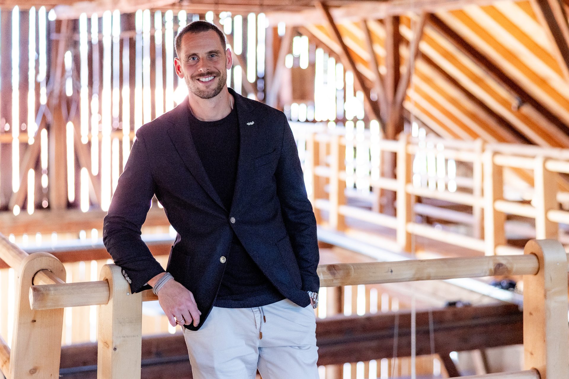 A man in a blazer and light pants smiles and leans against a wooden railing in a bright, airy space.