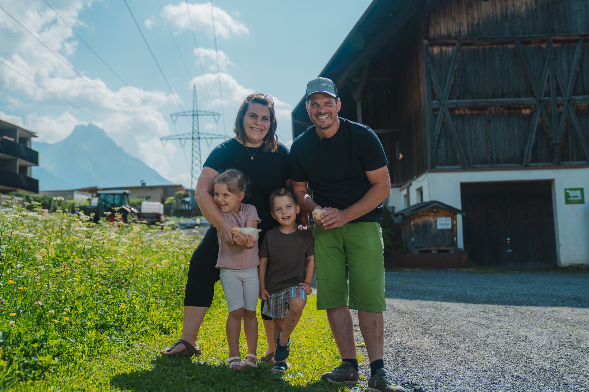 A smiling family of four, two adults and two children, holding baby chicks in a grassy field with mountains.