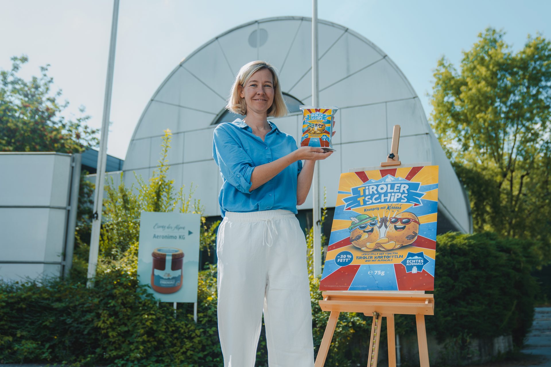 A woman presents Tiroler Tschips next to an easel displaying a vibrant chip poster in an outdoor setting.