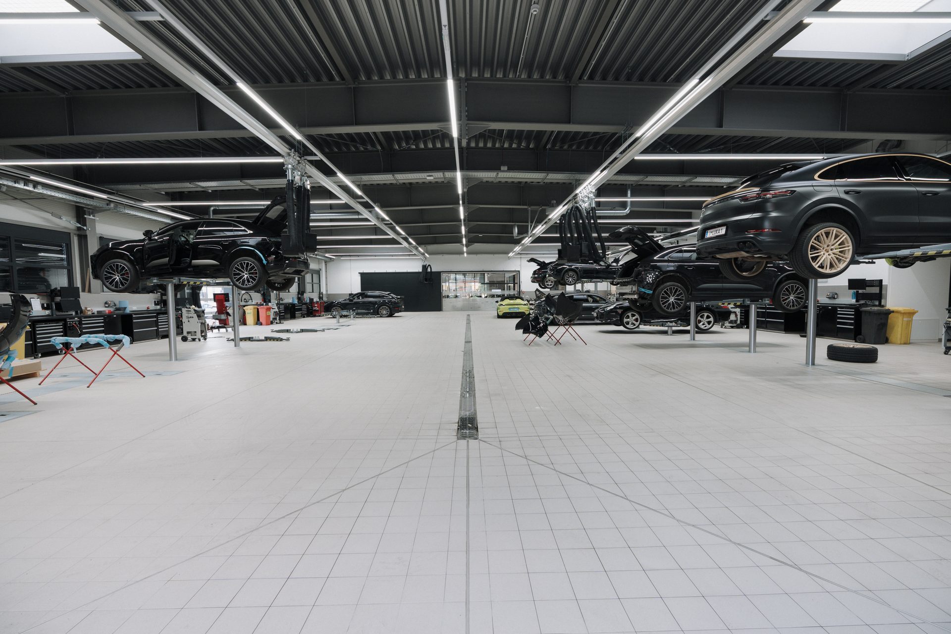 Porsche workshop with several cars, including SUVs, on lifts and parked on the floor, under bright lights.
