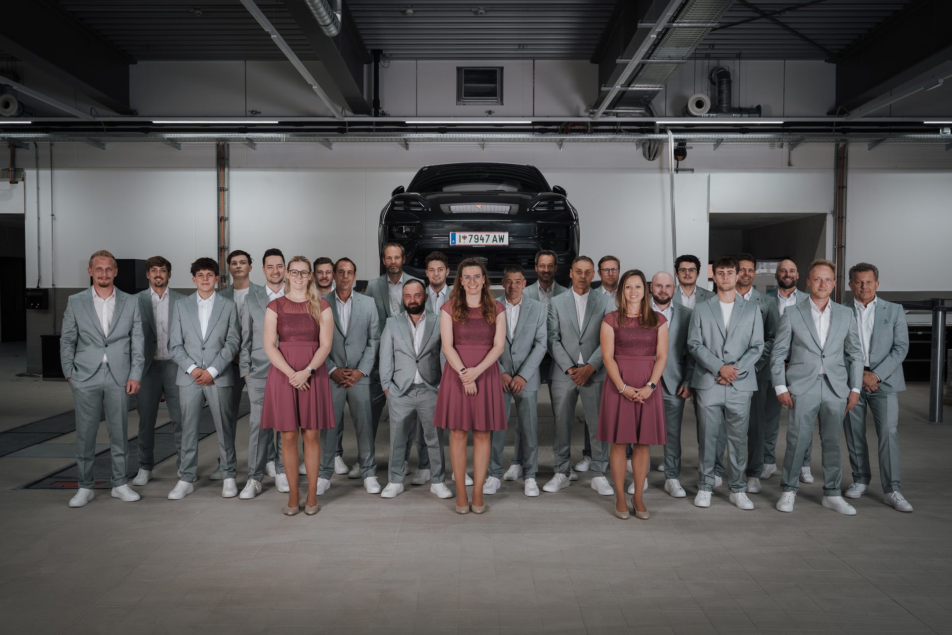 A team photo of men in grey suits and women in maroon dresses, posing in an automotive garage with a black car on a lift.