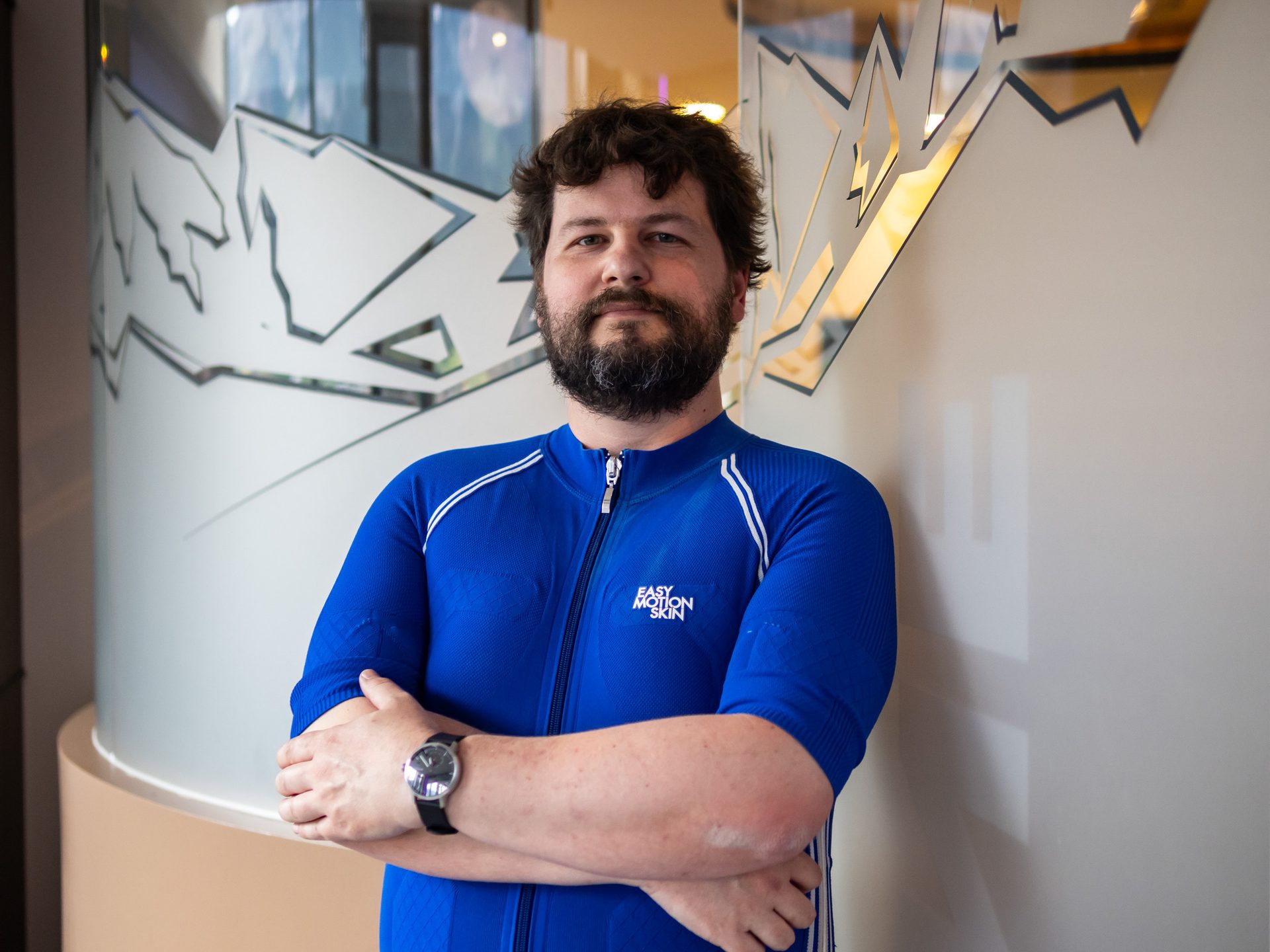 A man with a beard and curly hair, wearing a blue zip-up shirt, stands with arms crossed.