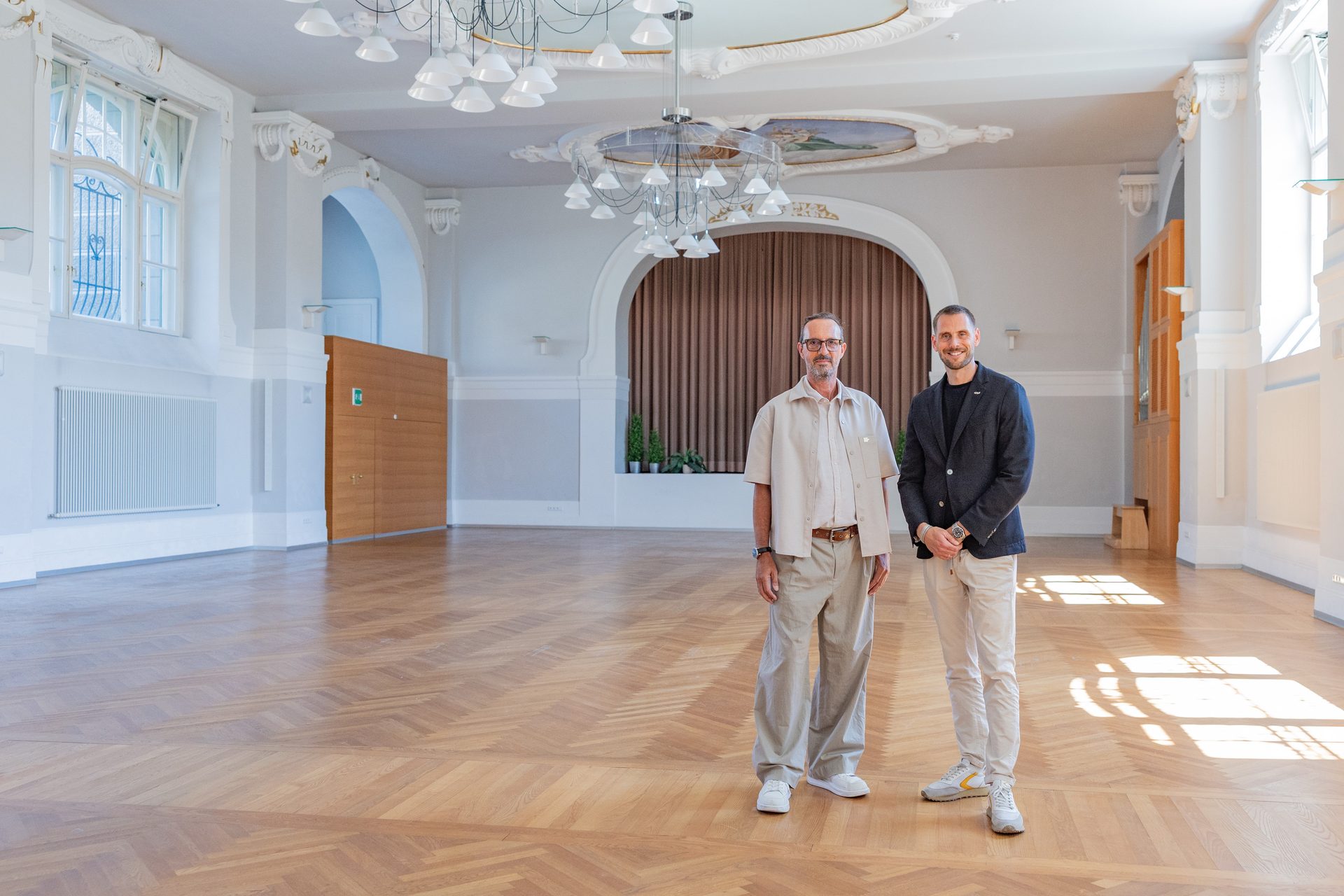 Two men stand smiling in a large, elegant hall with a parquet floor, ornate ceiling, and large windows.