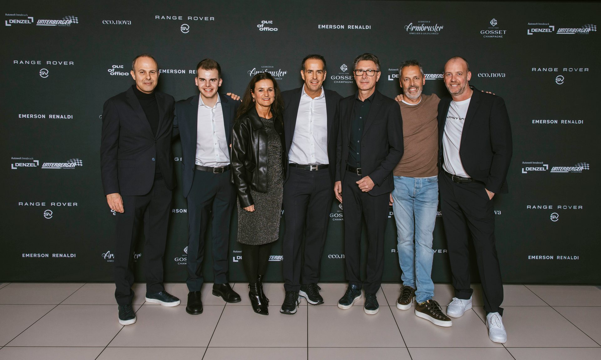 Seven people, including a woman, stand smiling in front of a dark wall adorned with company logos.